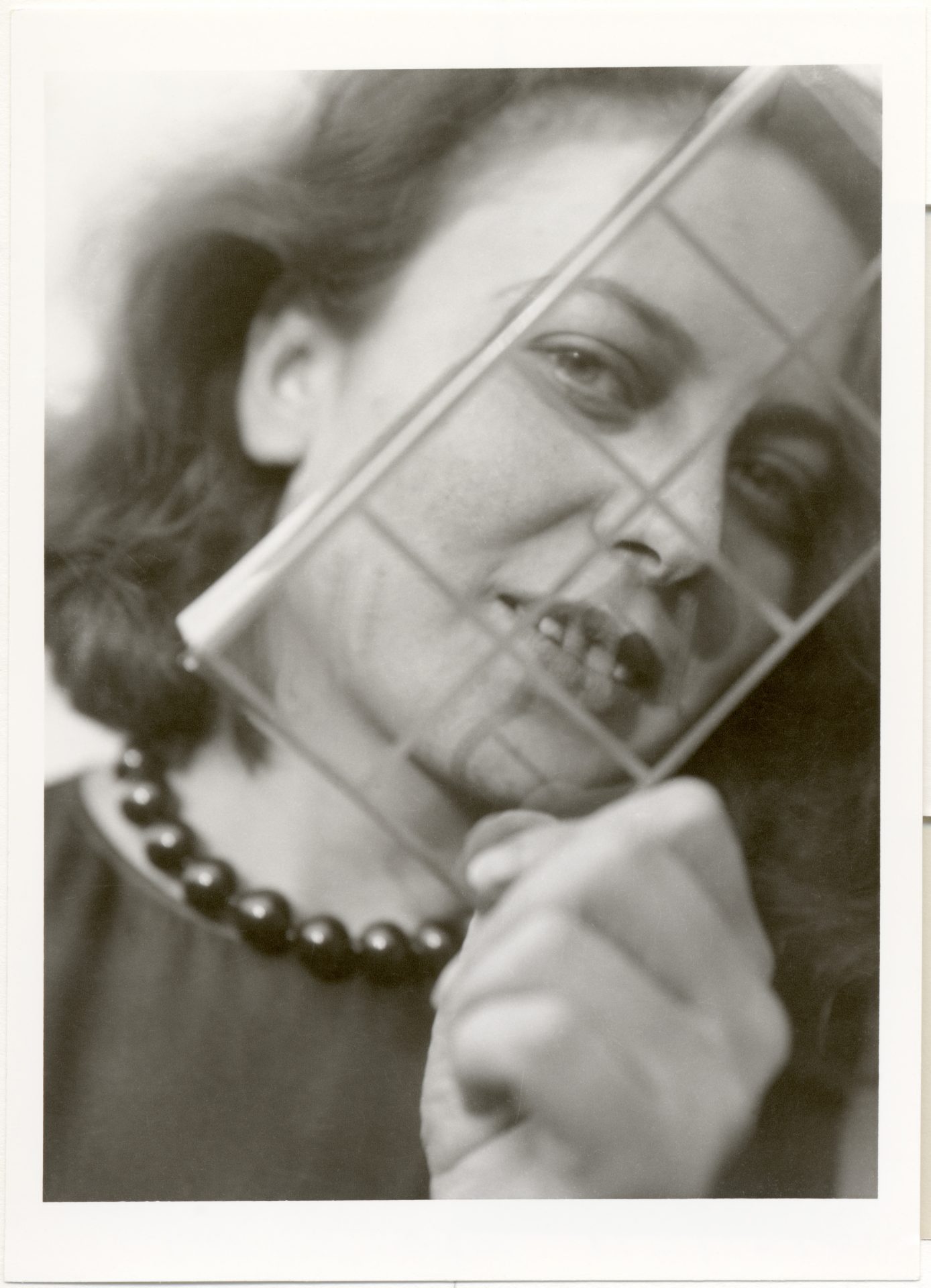 Black-and-white portrait of a smiling woman holding a lattice frame. She is wearing a pearl necklace.