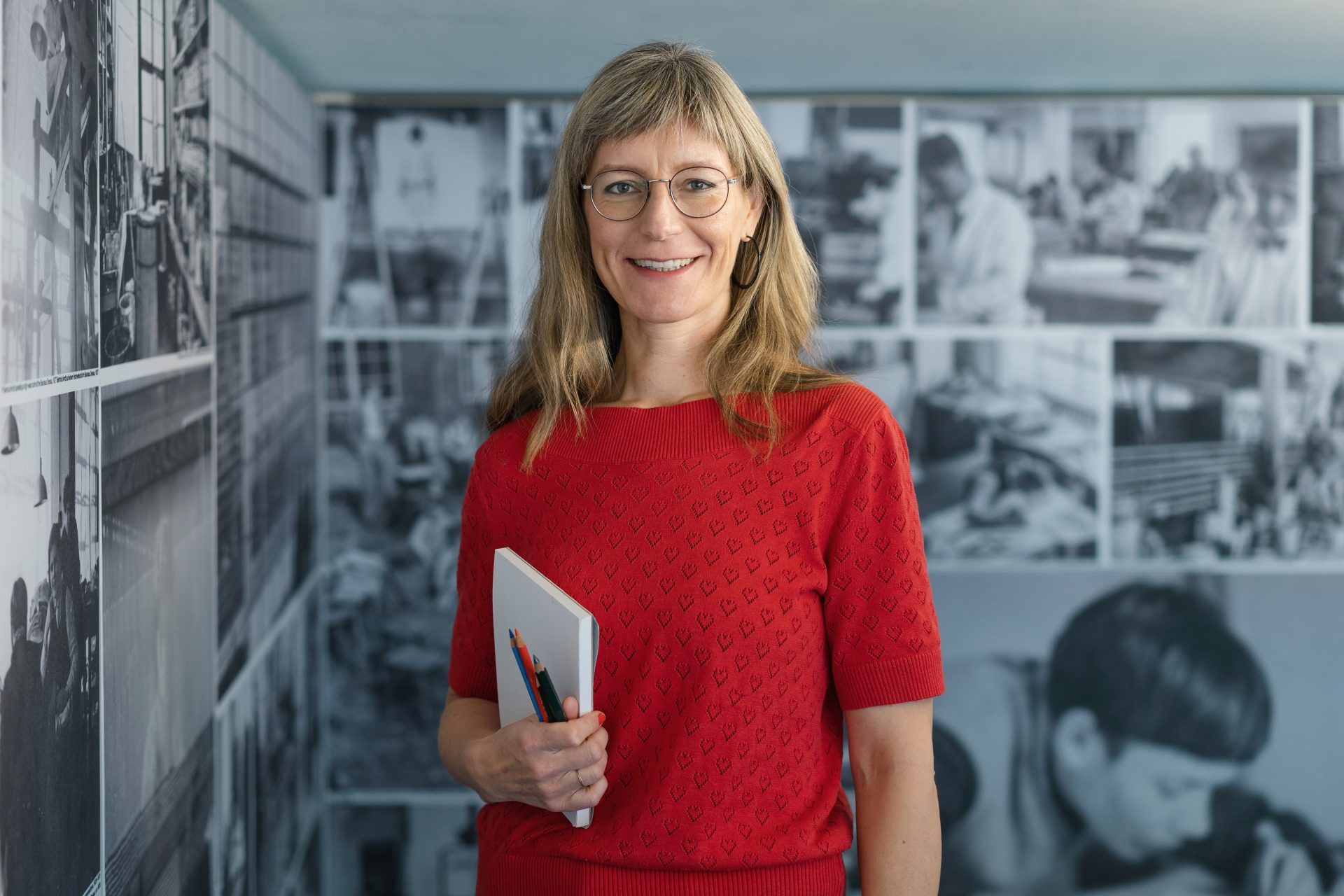 Foto einer Person mit Brille, langen blonden Haaren und rotem Oberteil, die ein Notizbuch vor einer Wand mit Schwarz-Weiß-Fotos hält.