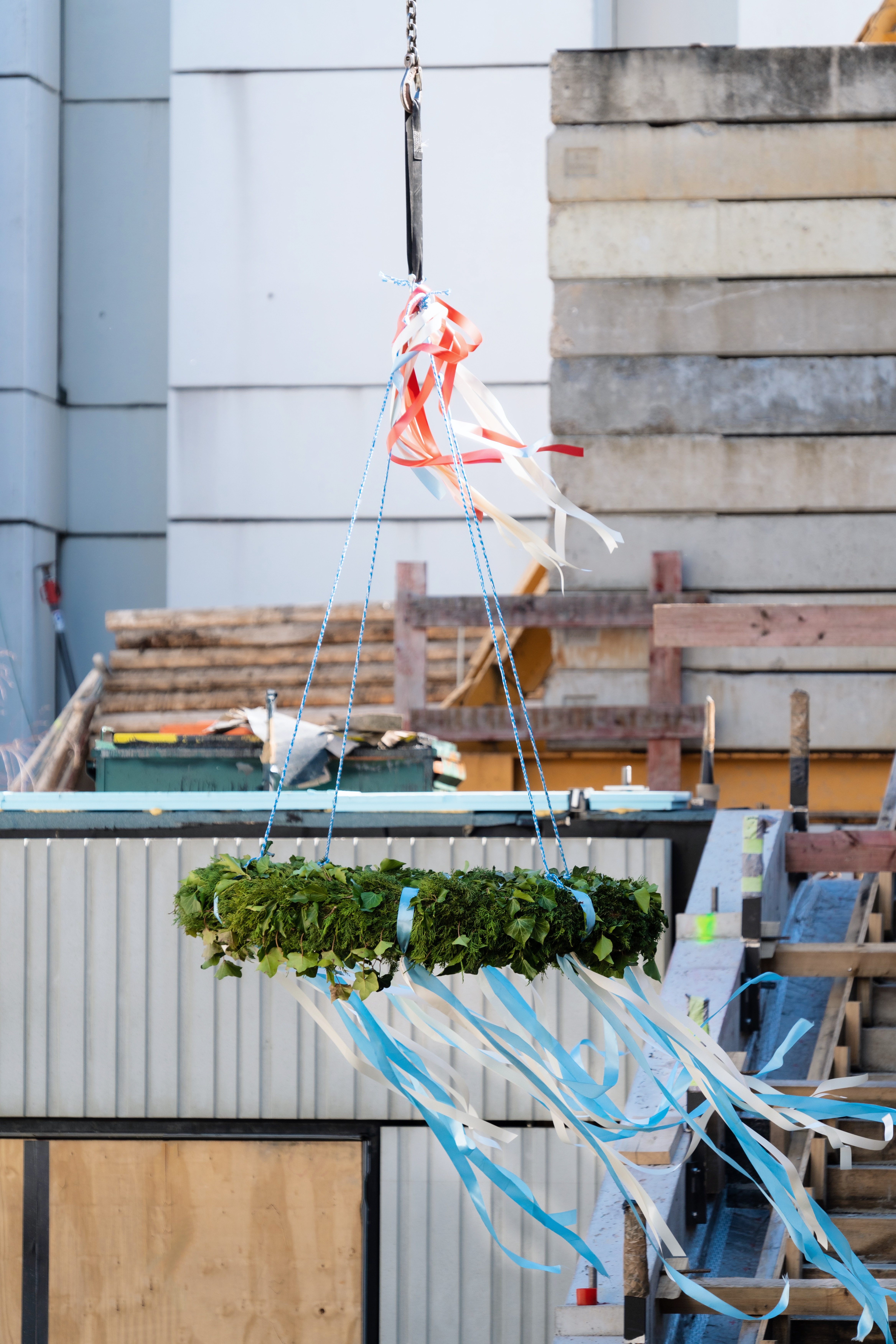 Ein hängender Kranz aus grünen Pflanzen, umgeben von bunten Bändern, schwebt über einer Baustelle.
