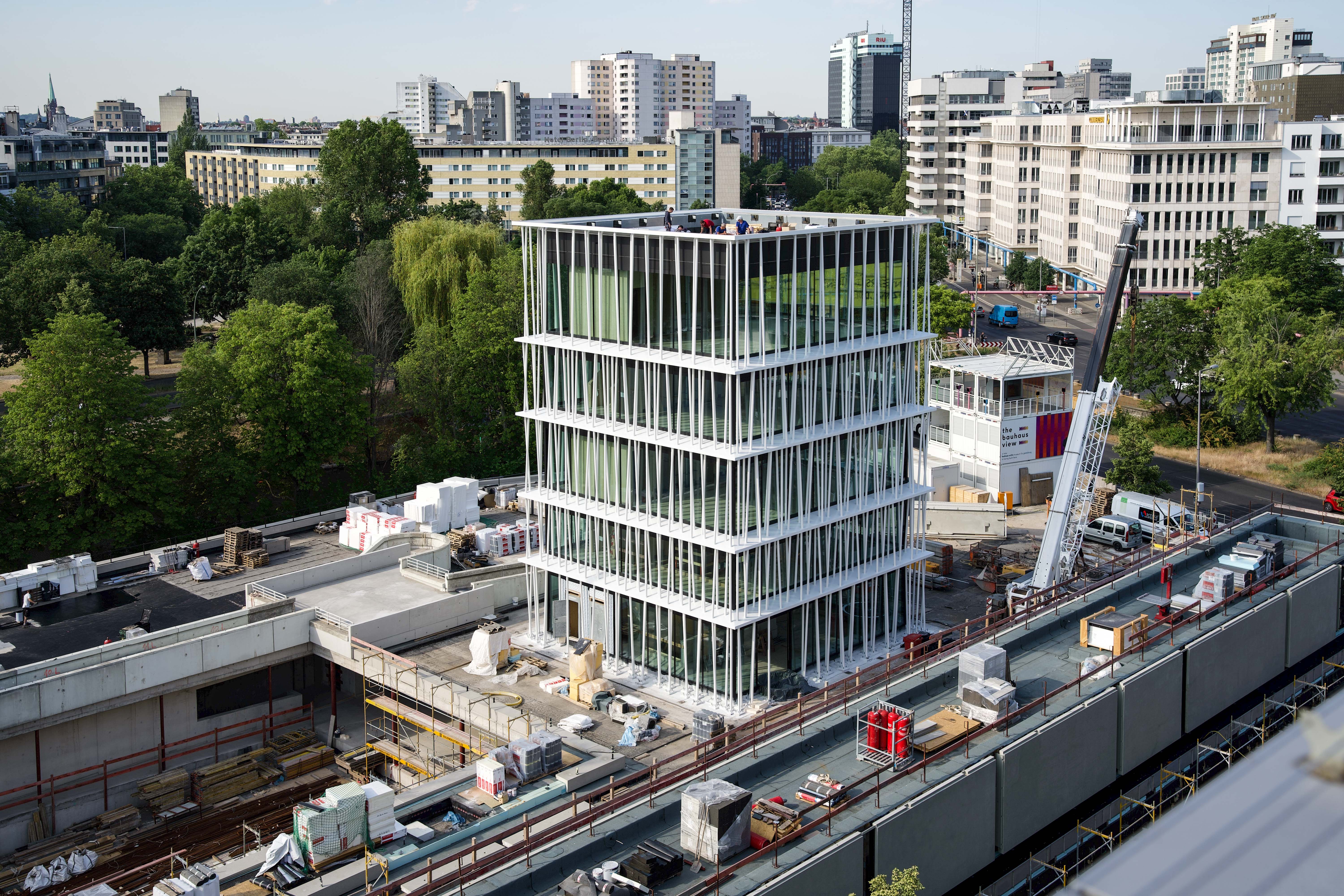 Moderne Baustelle mit gläsernem, mehrstöckigem Gebäude, umgeben von urbaner Landschaft und Bäumen.