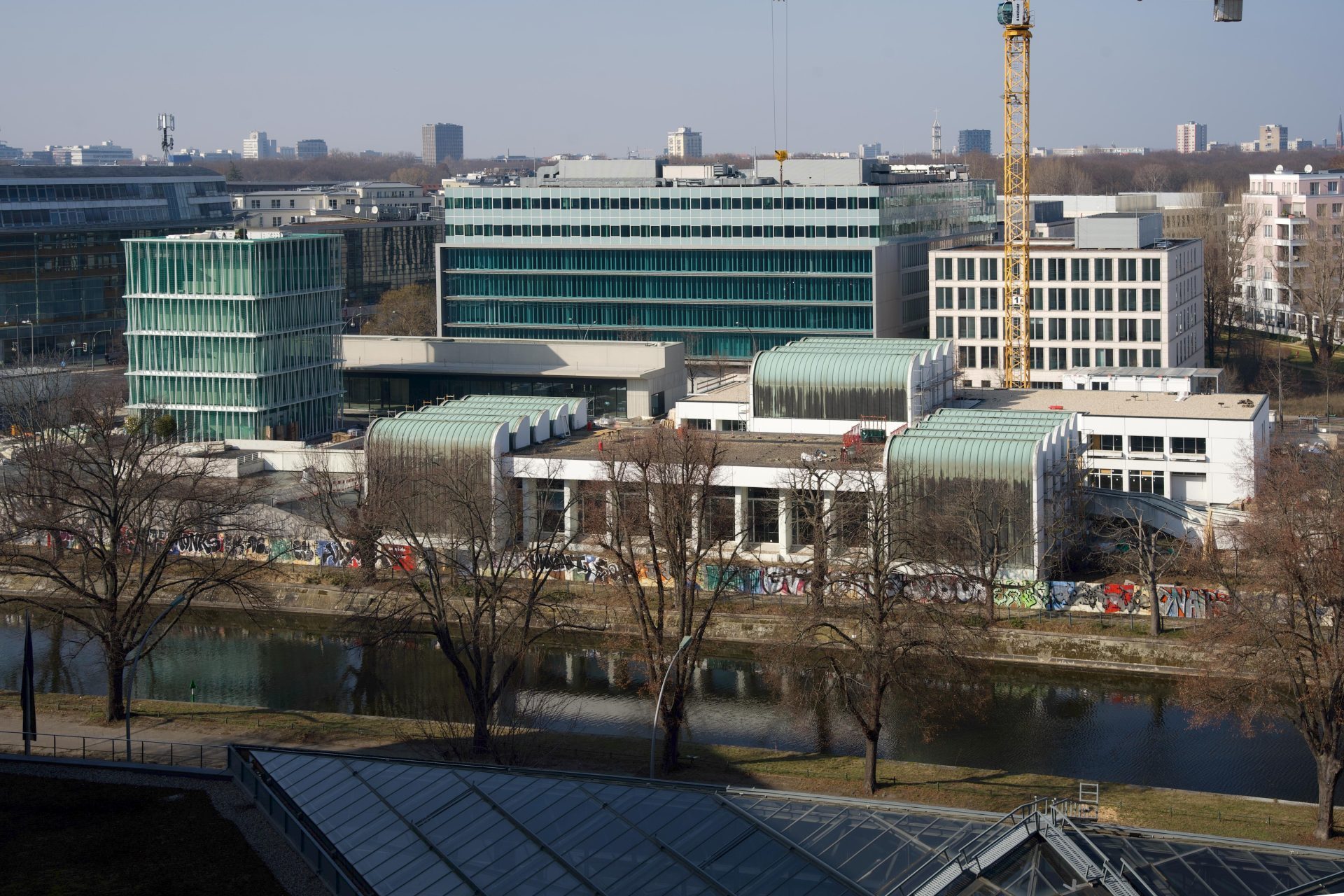 Modern buildings along a river, with trees in the foreground and a crane in the background.