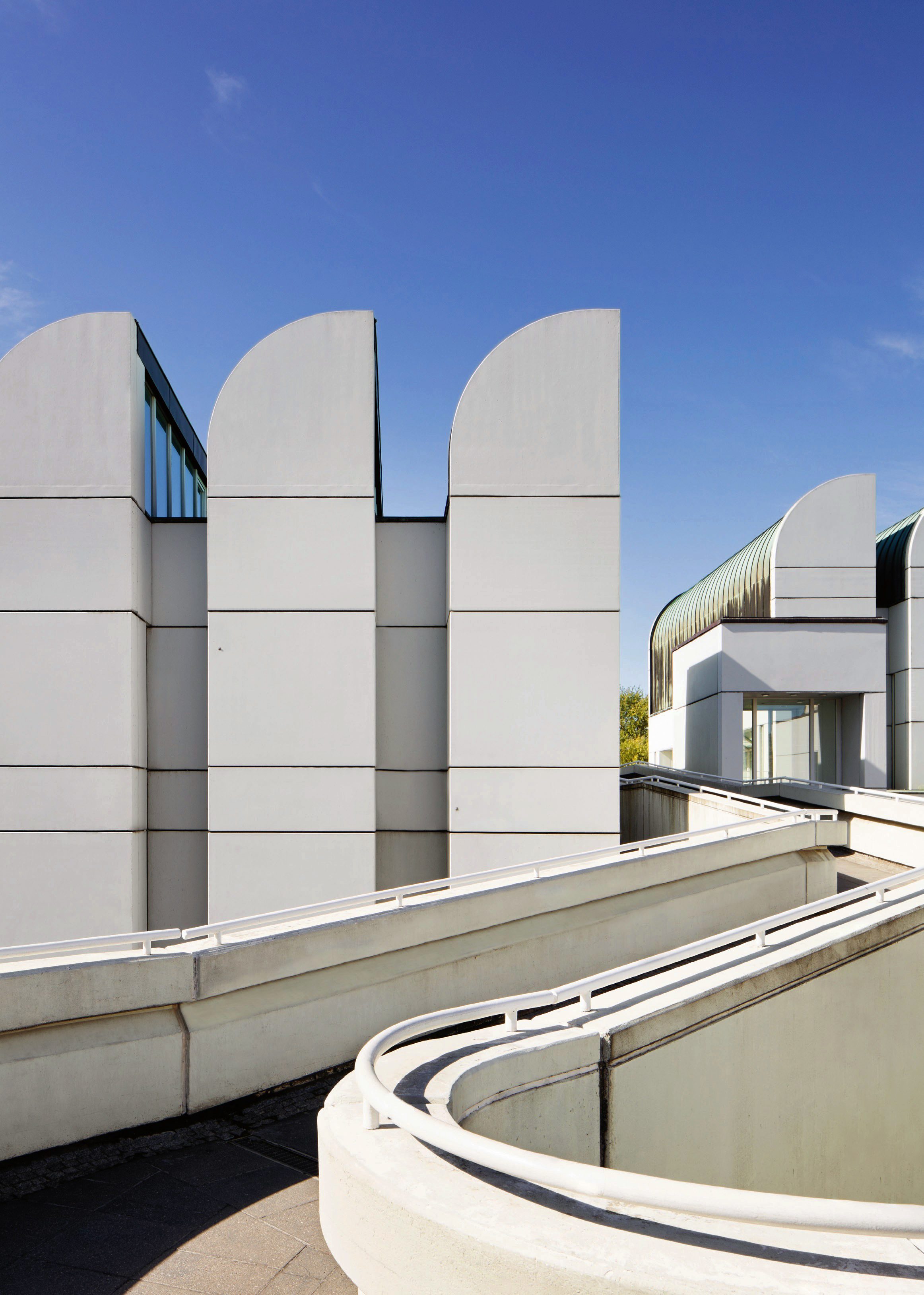 Foto des weißen Altbaus des Bauhaus-Archivs mit Shed-Dächern und geschwungener Rampe vor blauem Himmel.