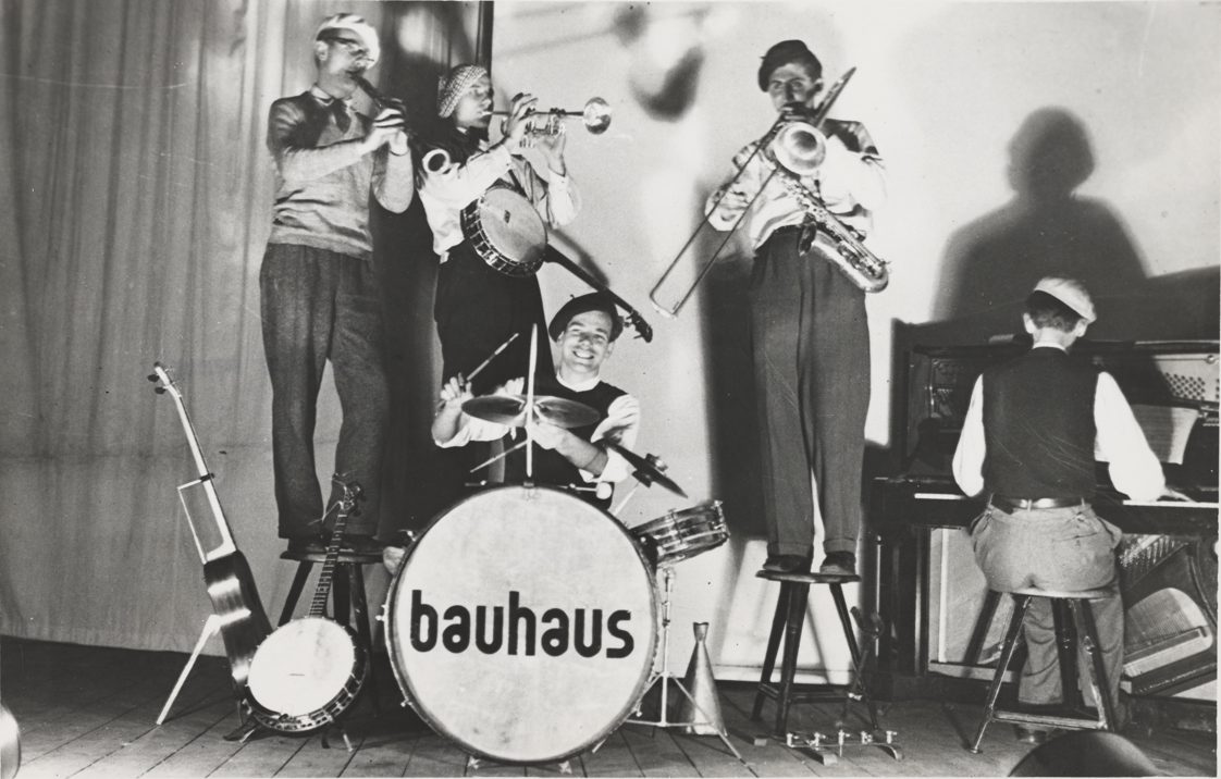 Bauhaus-Band mit vier Musikern auf der Bühne, darunter Trompete, Schlagzeug und Klavier.