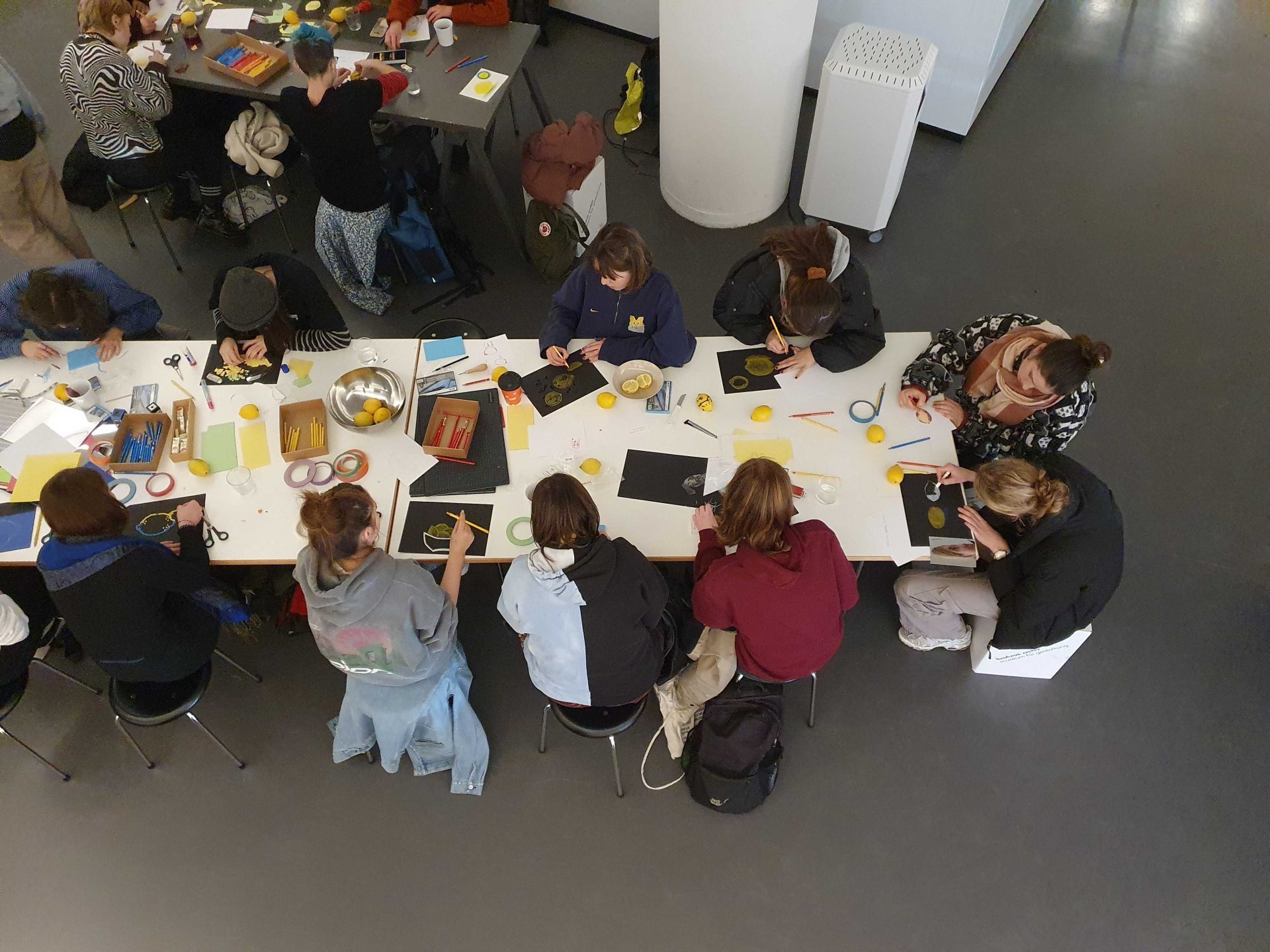 Foto einer Gruppe junger Menschen, mit mit Papier, Stiften und Zitronen an einem langen weißen Tisch sitzt und kümstlerisch arbeitet.