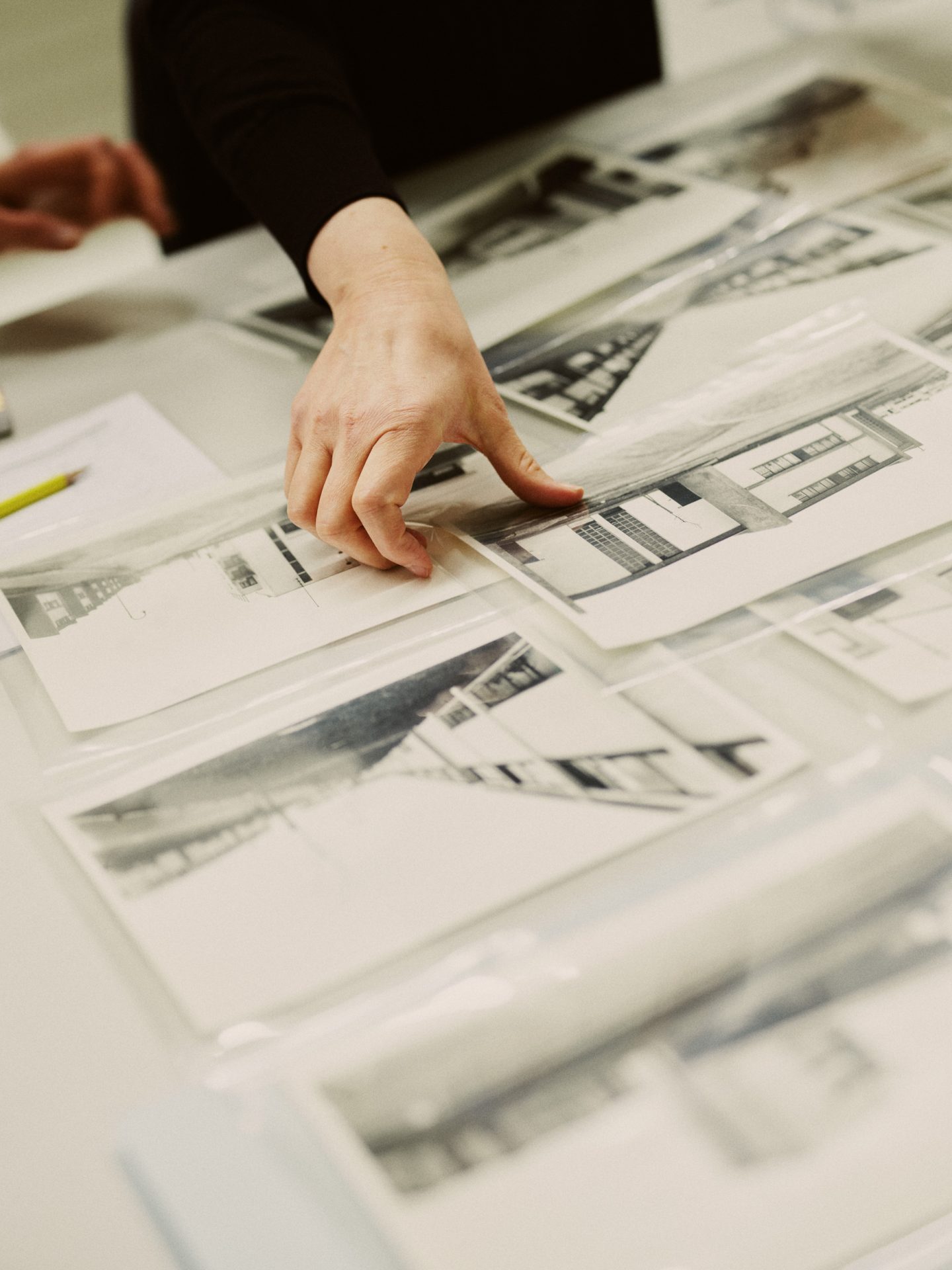 Foto eines Archivtischs, auf dem verschiedene Architekturfotografieren (schwarz-weiß) in durchsichtigen Hüllen ausgebreitet sind. Eine Hand greift nach einer Fotografie.
