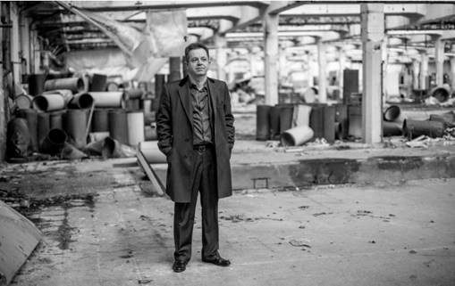Schwarz-Weiß-Foto eines Mannes im Anzug, der mit den Händen in den Taschen in einer verlassenen, industriellen Umgebung voller Rohre steht.