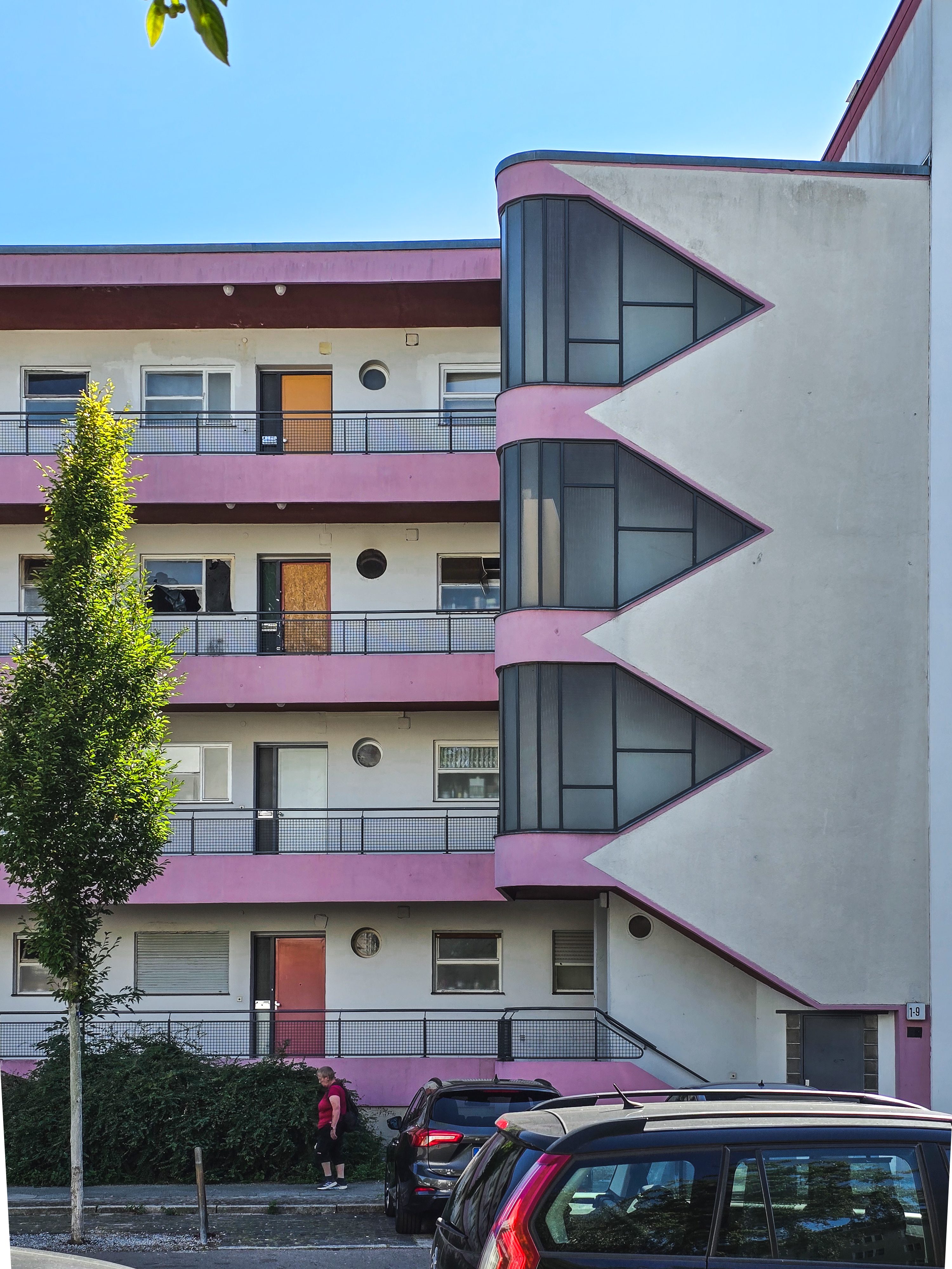 Modernes Wohngebäude mit pinken Akzenten, Glasfront und begrünten Parkplätzen unter blauem Himmel.