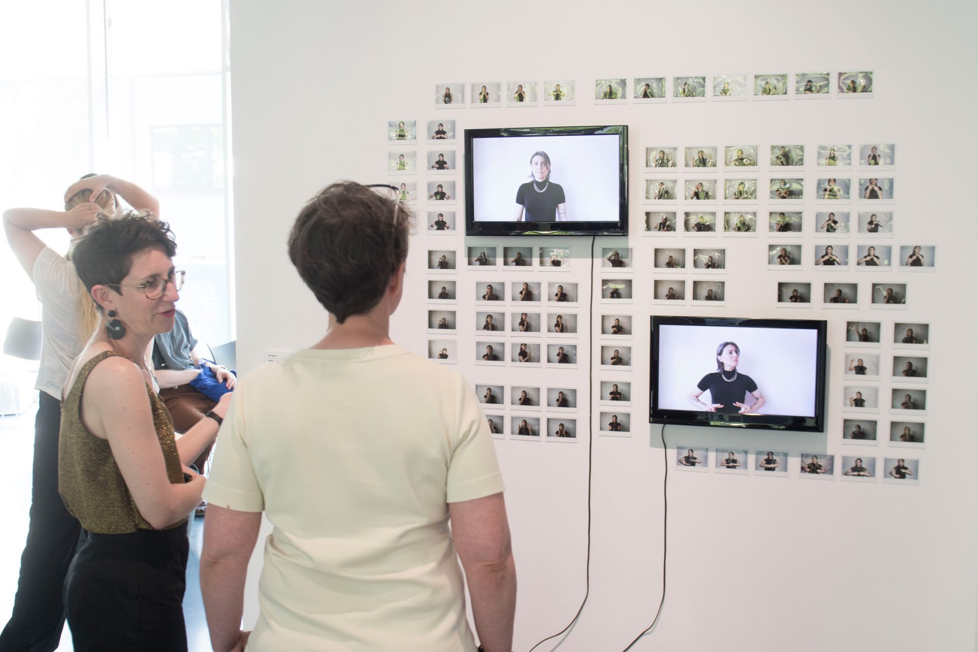 Drei Personen betrachten eine Wand mit zwei Bildschirmen und zahlreichen kleinen Fotos, auf denen Gebärdensprache demonstriert wird.
