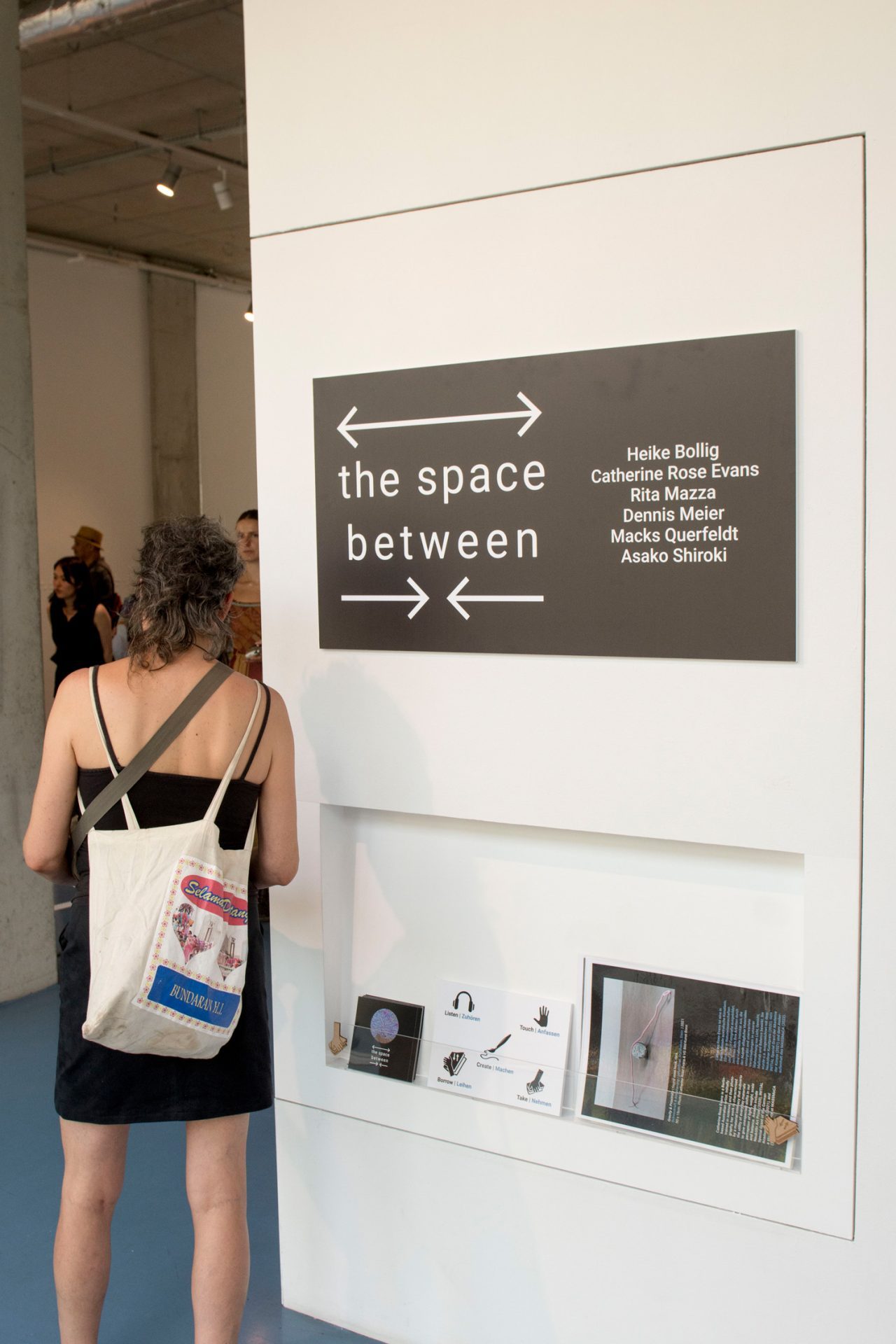 Eine Person steht im Ausstellungsraum vor einem schwarzen Schild mit weißer Schrift, auf dem der Titel der Ausstellung und die Namen der Künstler und Künstlerinnen stehen.