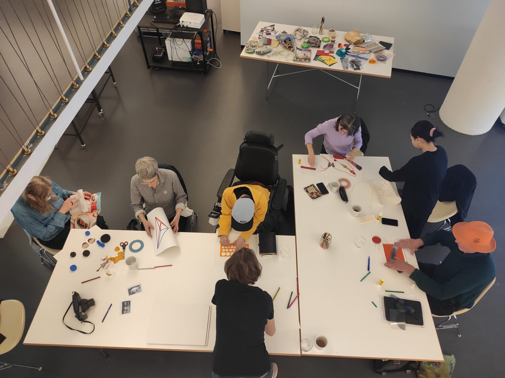 Sieben Personen sitzen an zwei zusammengestellten Tischen und arbeiten gemeinsam an kreativen Projekten mit Papier, Stiften und Bastelmaterialien.