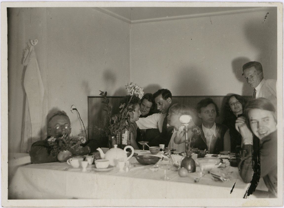Group scene with six people at a table set with dishes and a teapot, in a simple room.