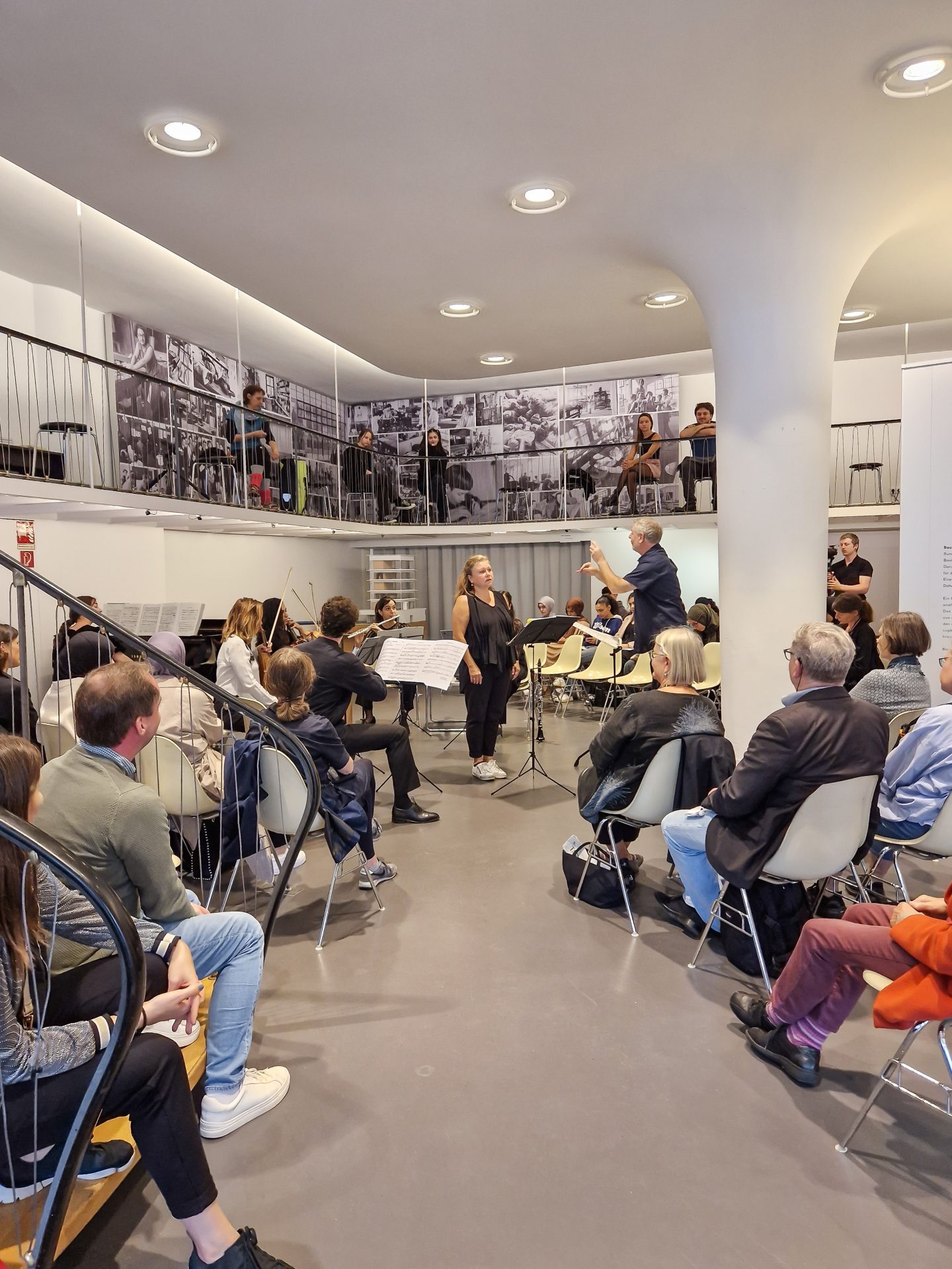 Rehearsal with a conductor and an orchestra, surrounded by spectators in a modern space.