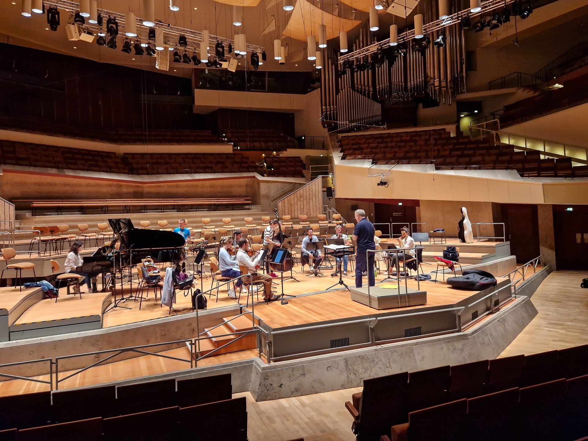 Orchestra rehearsing on stage with a piano and several musicians in a modern hall.