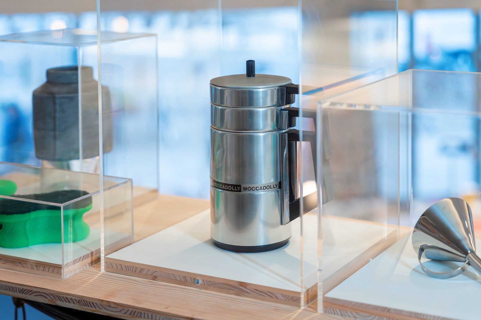 Edelstahl-Kaffeekanne in einer Vitrine, umgeben von weiteren Objekten wie einer grünen Skulptur und einem Trichter.