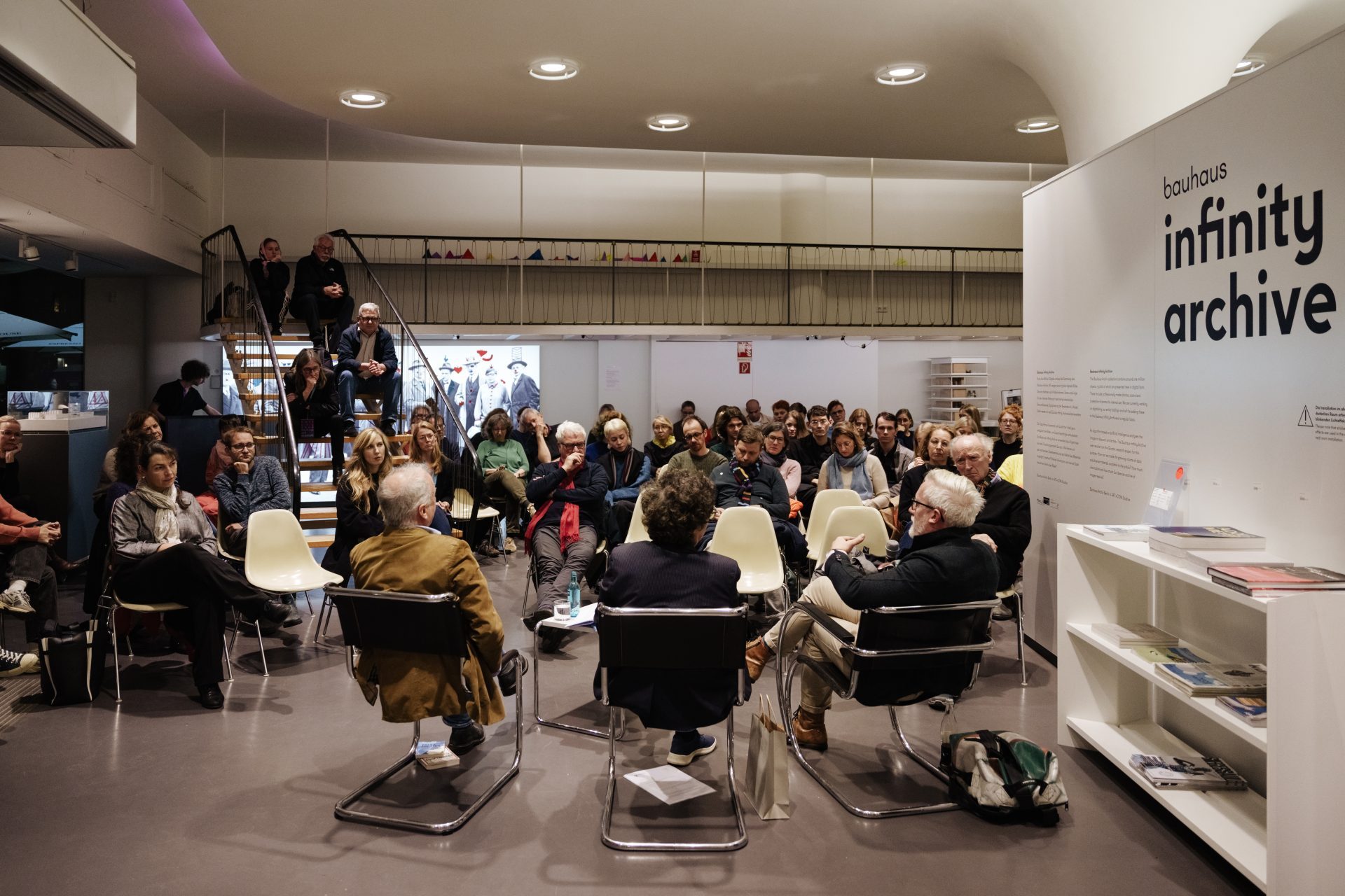 Diskussionsrunde mit mehreren Personen in modernem Raum, Publikum auf Stühlen, an der Wand steht "infinity archive".