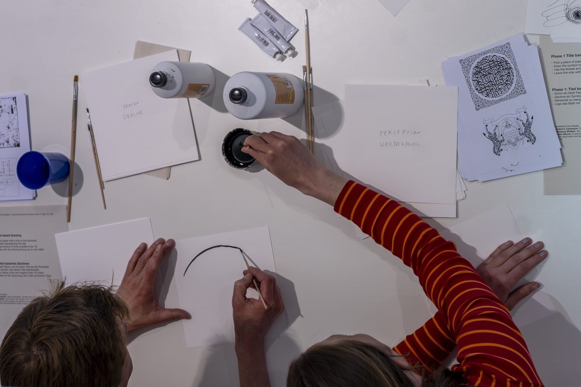 Zwei Personen zeichnen auf Papier, umgeben von Stiften, Notizen und anderen Materialien auf einem Tisch.