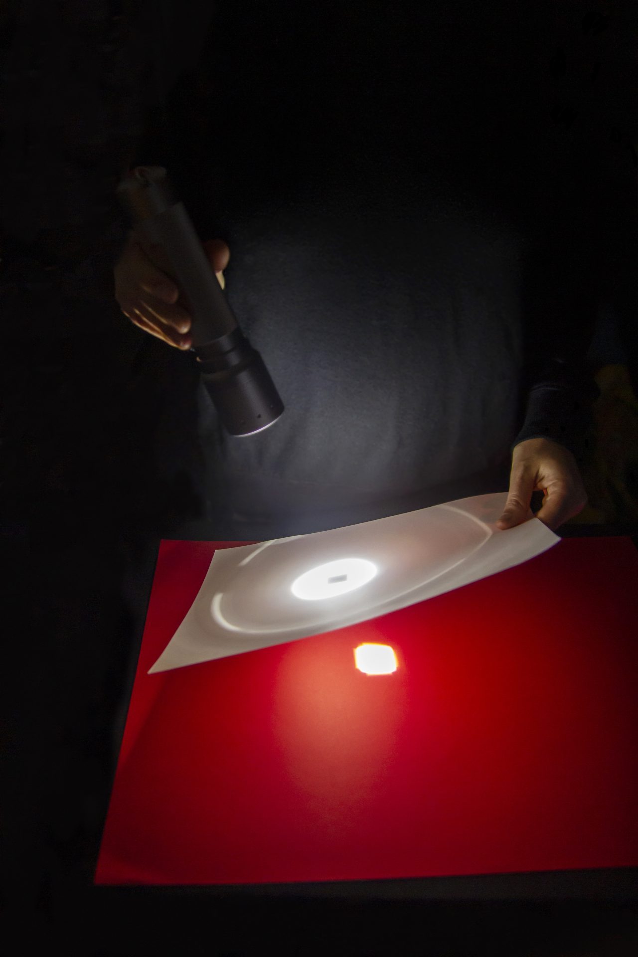 Hände halten eine Taschenlampe und einen transparenten Film vor einem roten Hintergrund, der Licht reflektiert.