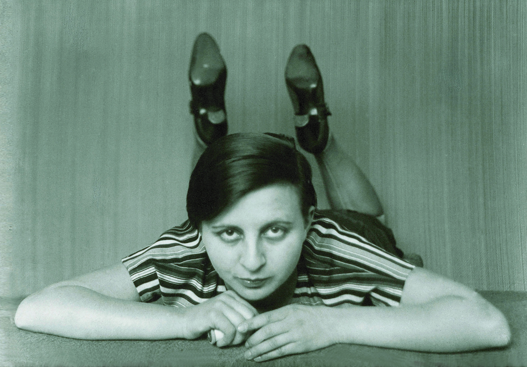 A woman in a striped outfit lies with supported hands and looks at the camera, shoes visible. Black and white photography.
