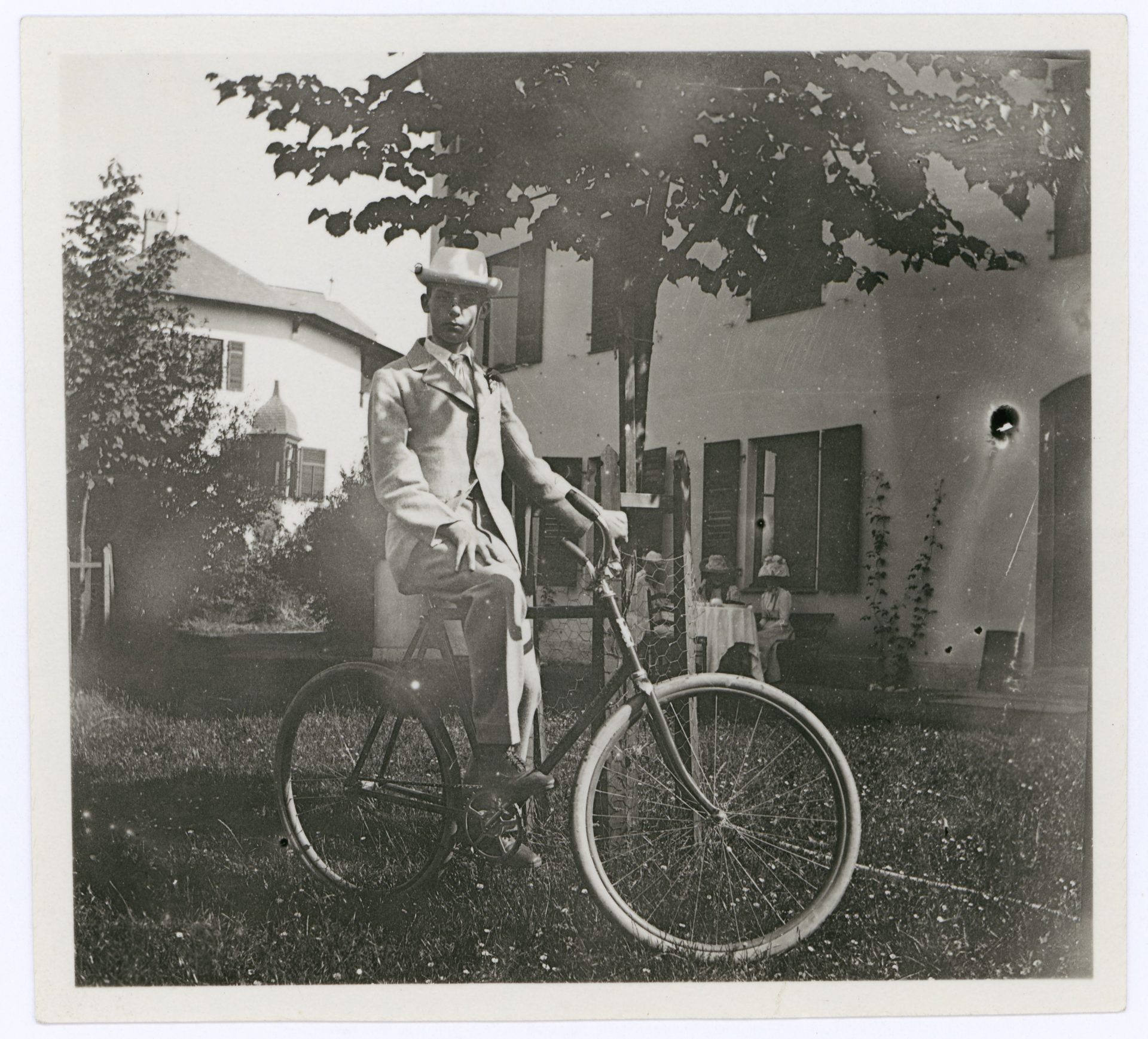 A man in a suit sits on an old bicycle in front of a house with trees and a seated person in the background.