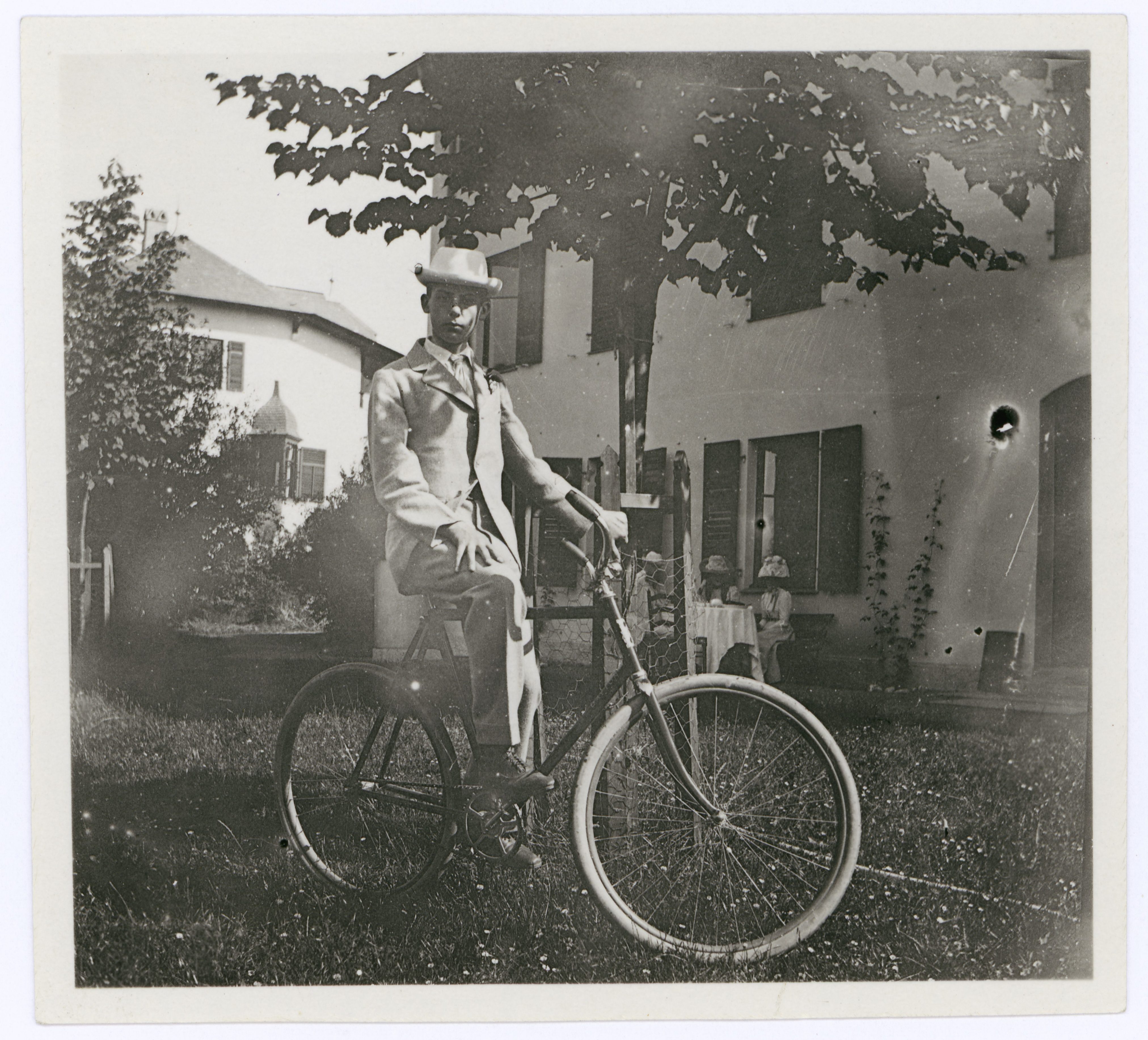 Ein junger Mann im Anzug sitzt auf einem alten Fahrrad vor einem Haus mit Bäumen und drei sitzenden Personen im Hintergrund.
