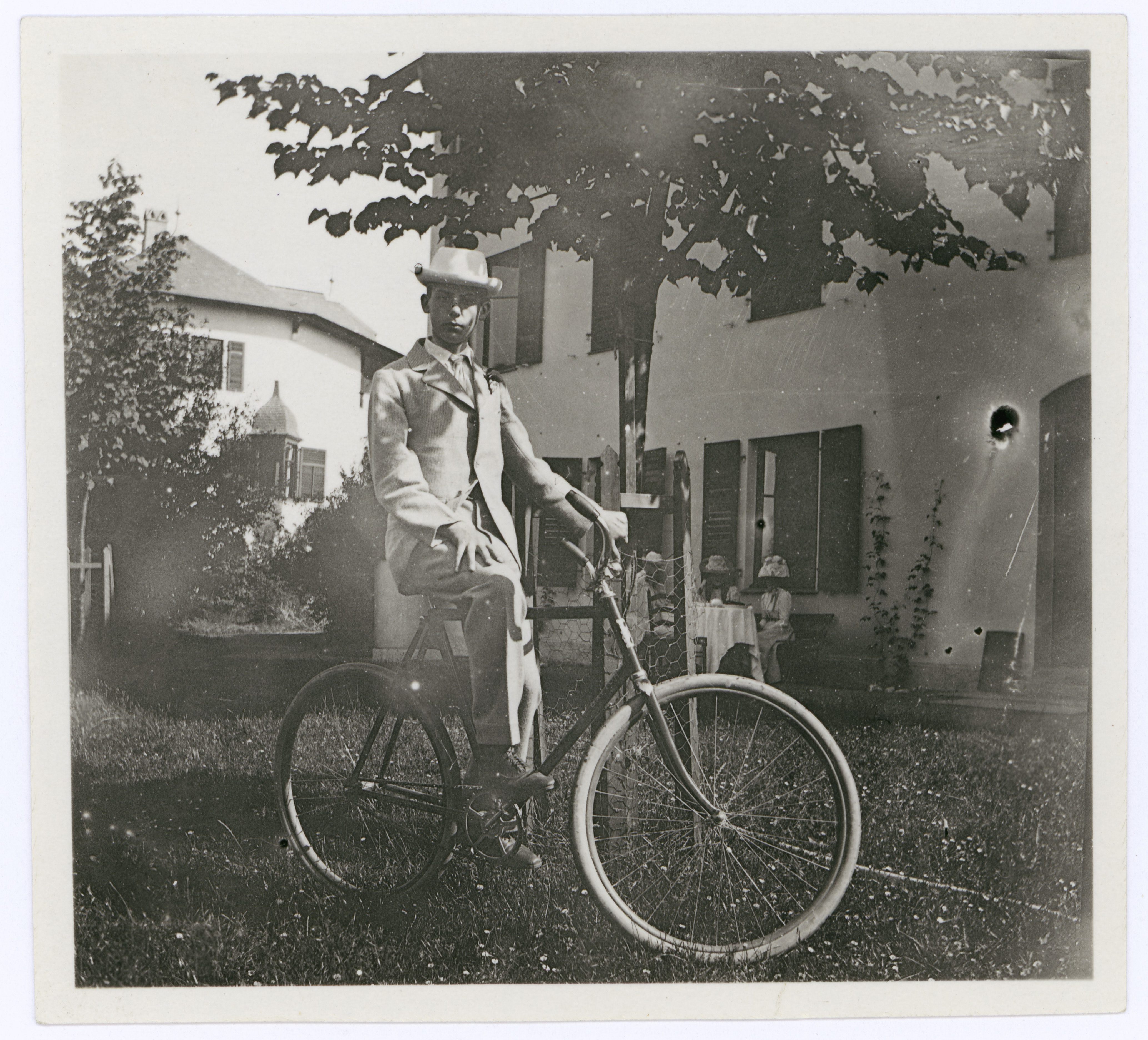 A man in a suit sits on an old bicycle in front of a house with trees and a seated person in the background.