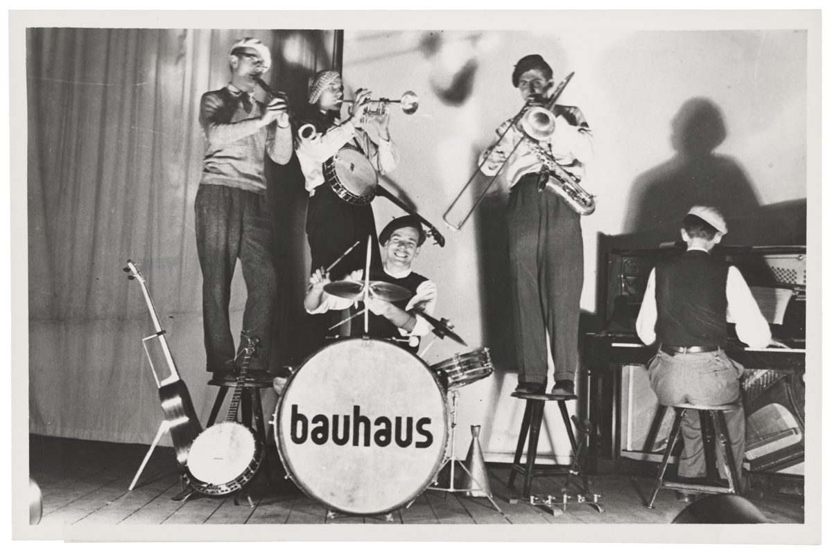Schwarz-weißes Bild einer Band mit verschiedenen Instrumenten auf der Bühne, "bauhaus"-Schriftzug auf dem Schlagzeug.