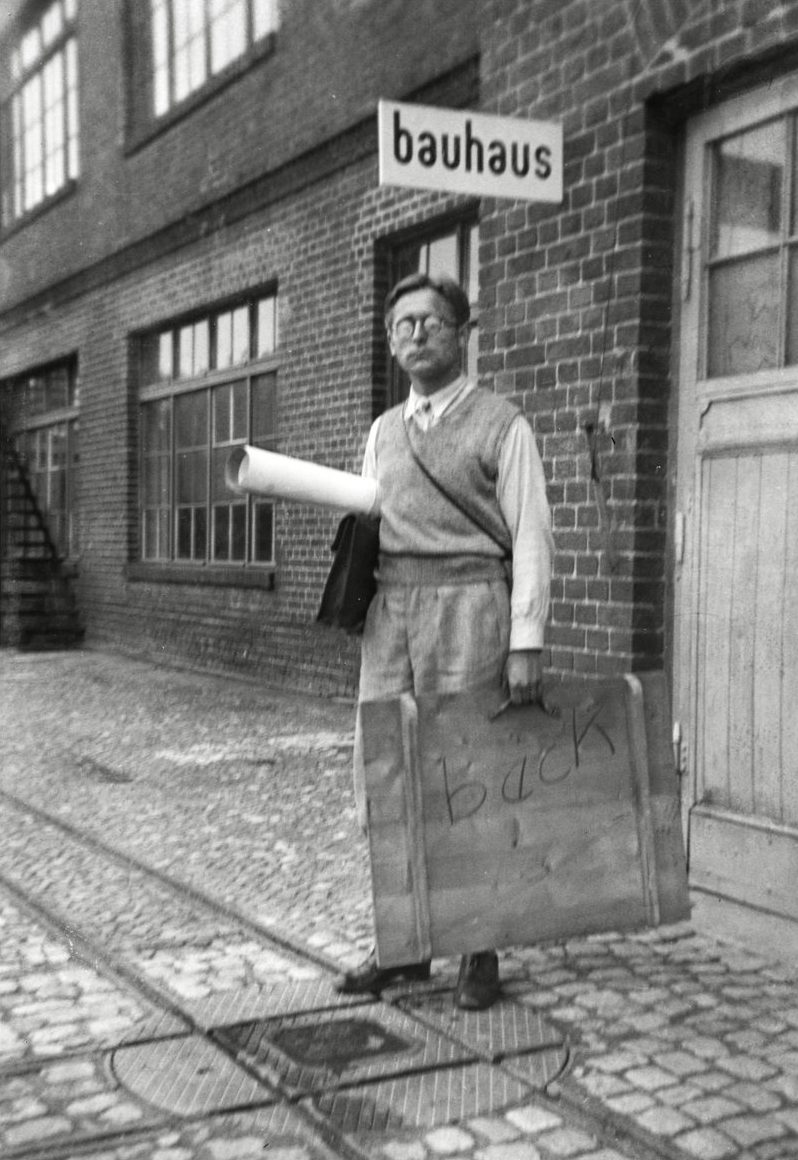 Man in front of the Bauhaus building holding a sign, carrying a design under his arm, historical black-and-white photograph.