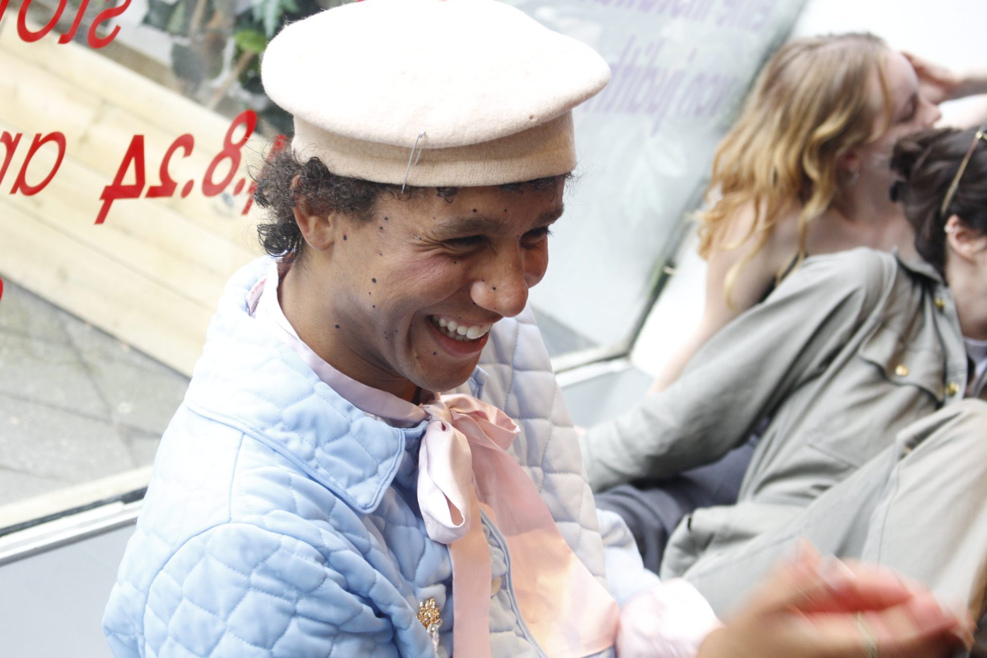 Smiling person in a blue jacket and pink bow, sitting in a café with other people in the background.