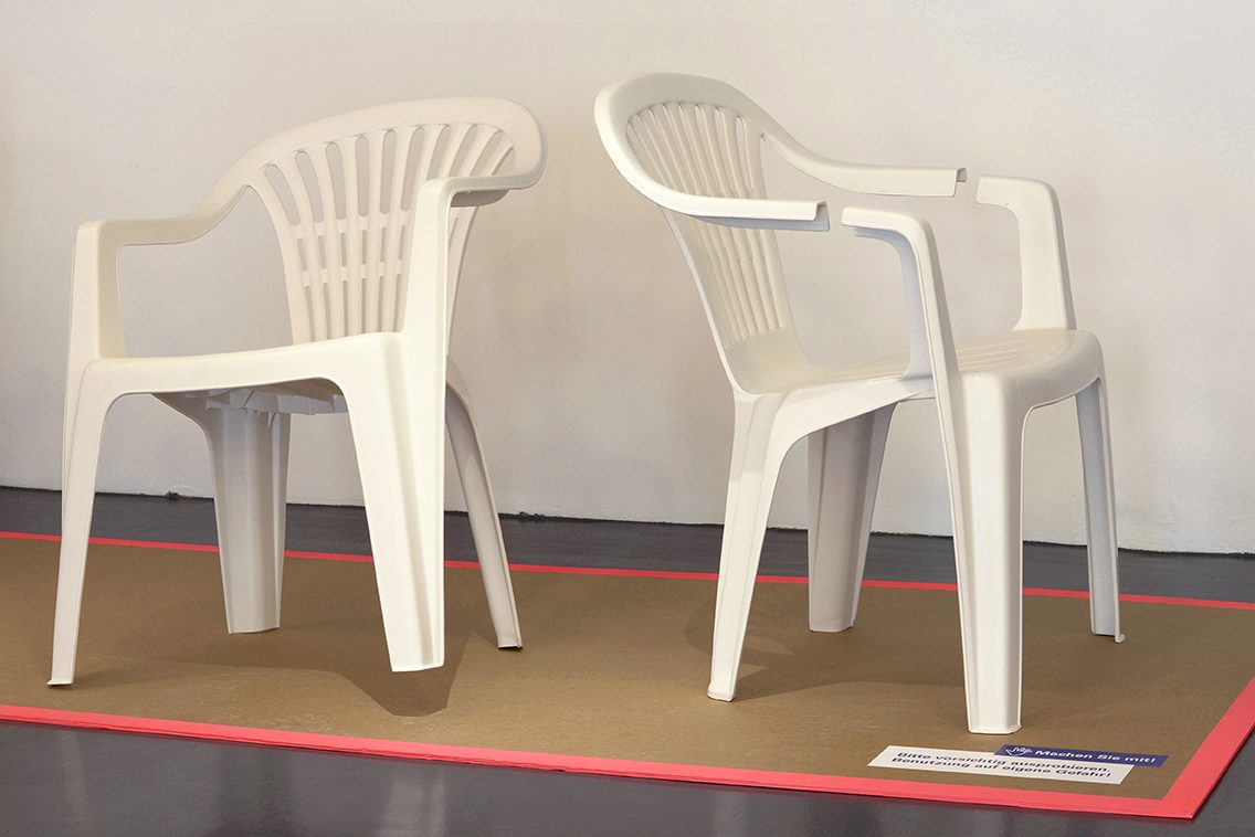 Zwei weiße Plastikstühle stehen auf einem Boden mit brauner Unterlage und pinkem Rand.