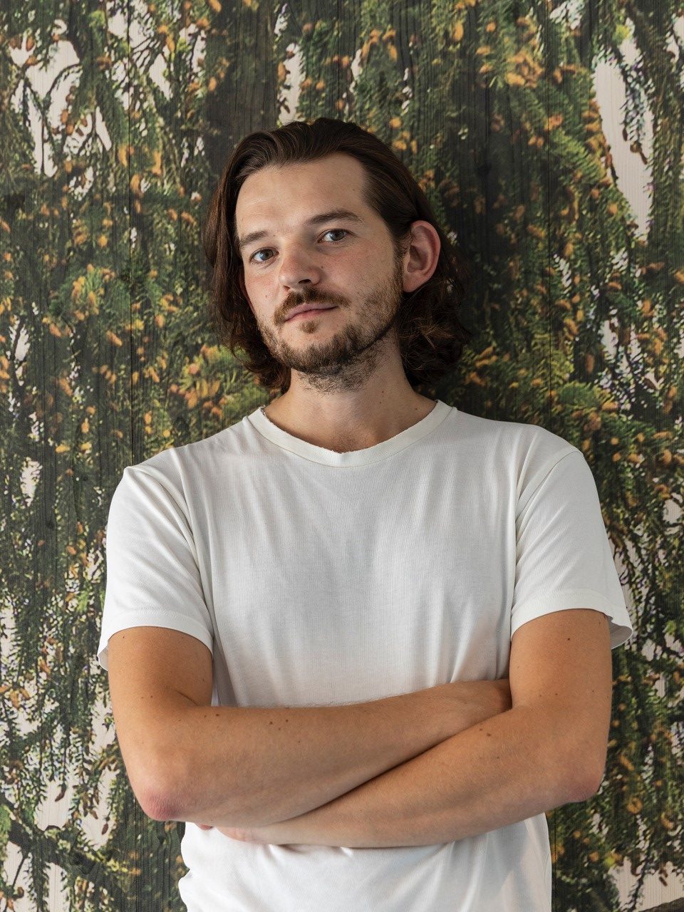 Mann mit dunklem Haar in weißem T-Shirt vor einer grünen Hintergrund, mit verschränkten Armen.