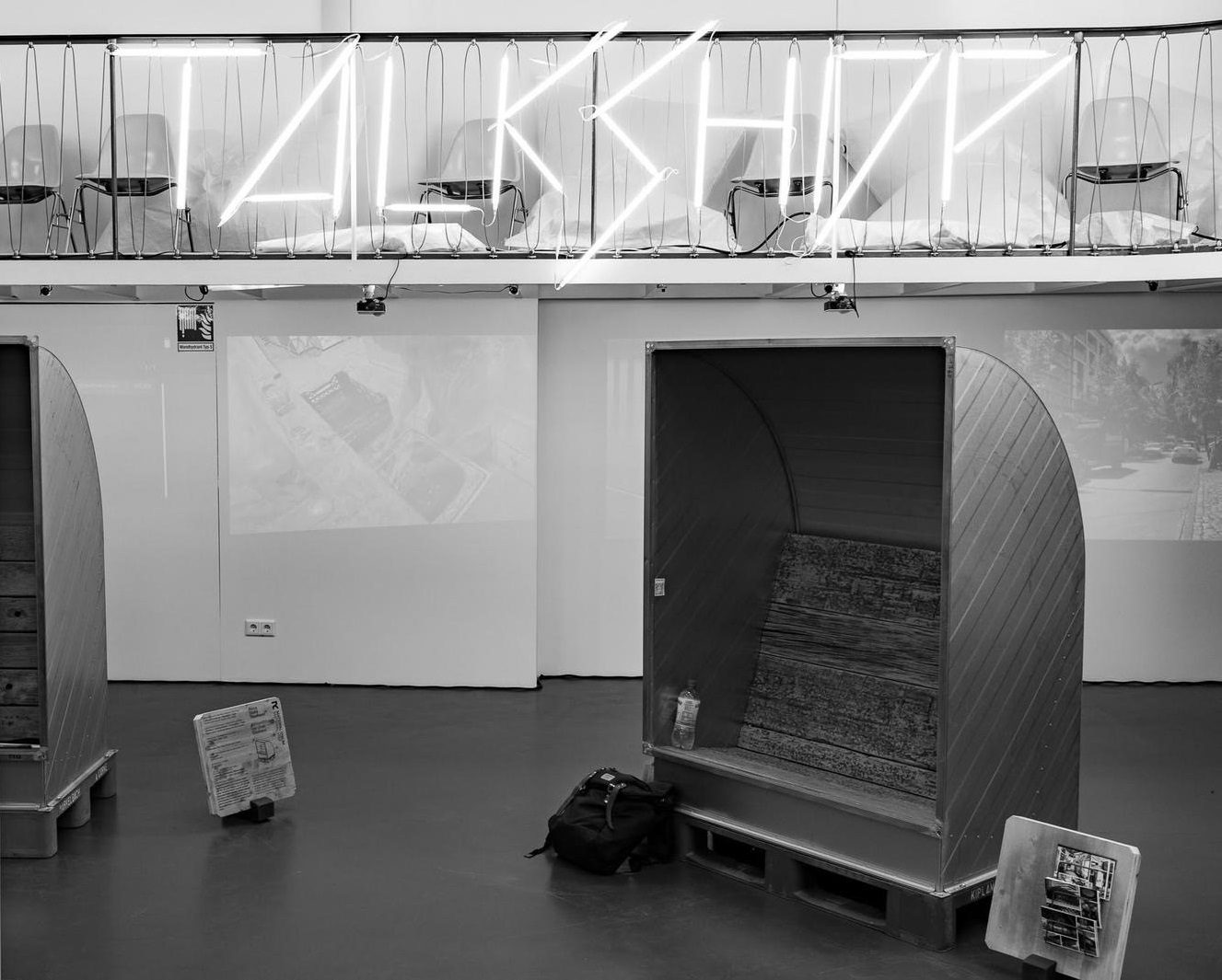 Moderne Galerie mit neonfarbenem "TALKSHOP"-Schild, Holzstrukturen und Hintergrundprojektionen.