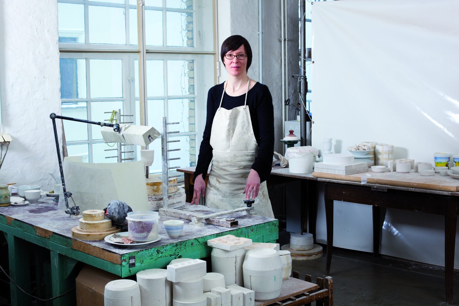 Künstlerin in einem Atelier mit Töpferwaren, Arbeitsfläche und Beleuchtung im Hintergrund.