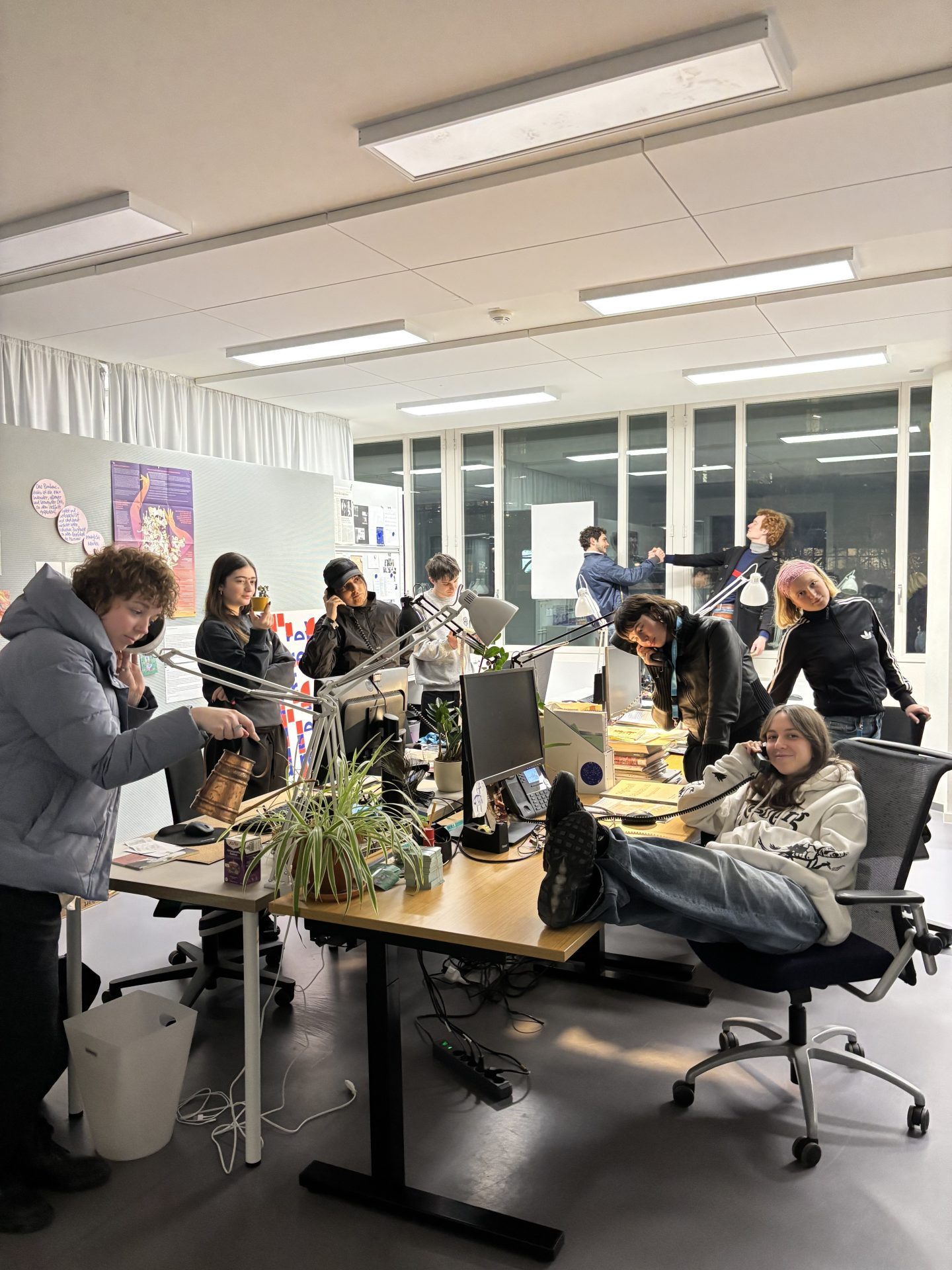 Group of people in a modern space, working on computers and chatting, surrounded by plants and books.