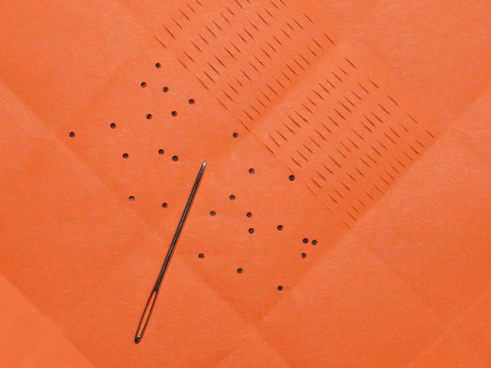Eine Nadel liegt auf orangefarbenem Papier, umgeben von kleinen Löchern und gestickten Linien.