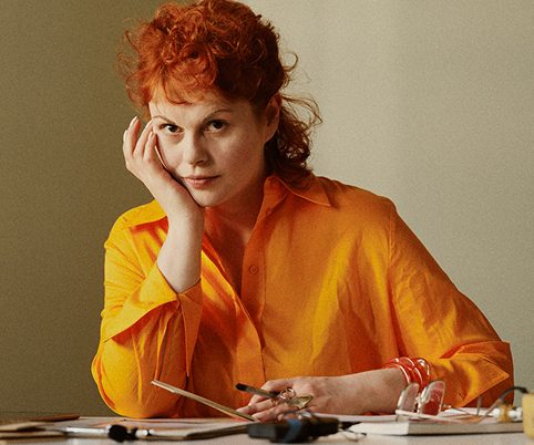 Frau mit rotem Haar in orangefarbener Bluse, die nachdenklich an einem Tisch sitzt. Stifte und Papiere liegen vor ihr.