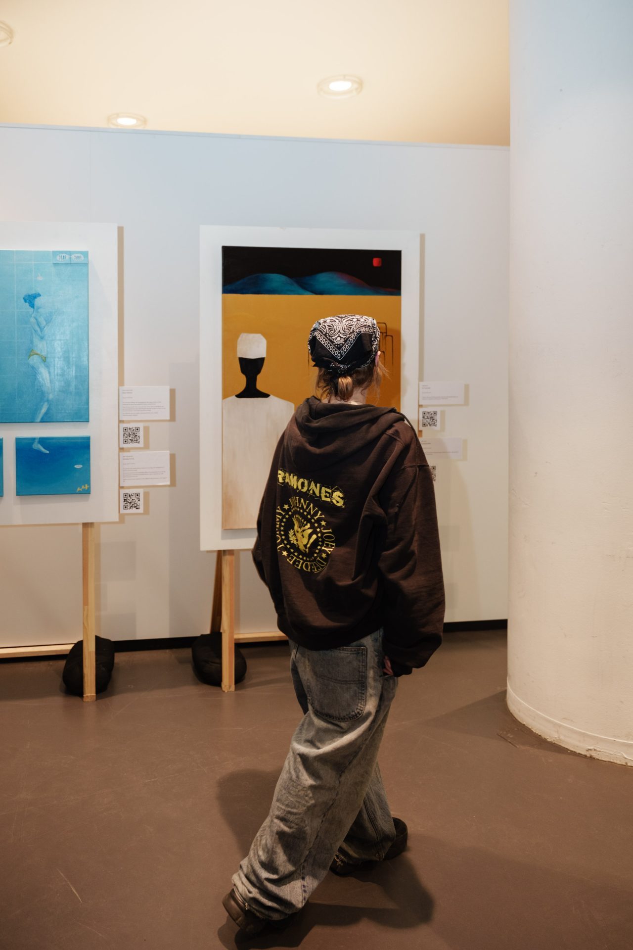 Person in Hoodie und weiten Hosen, die Kunstwerke in einem Ausstellungsraum betrachtet.
