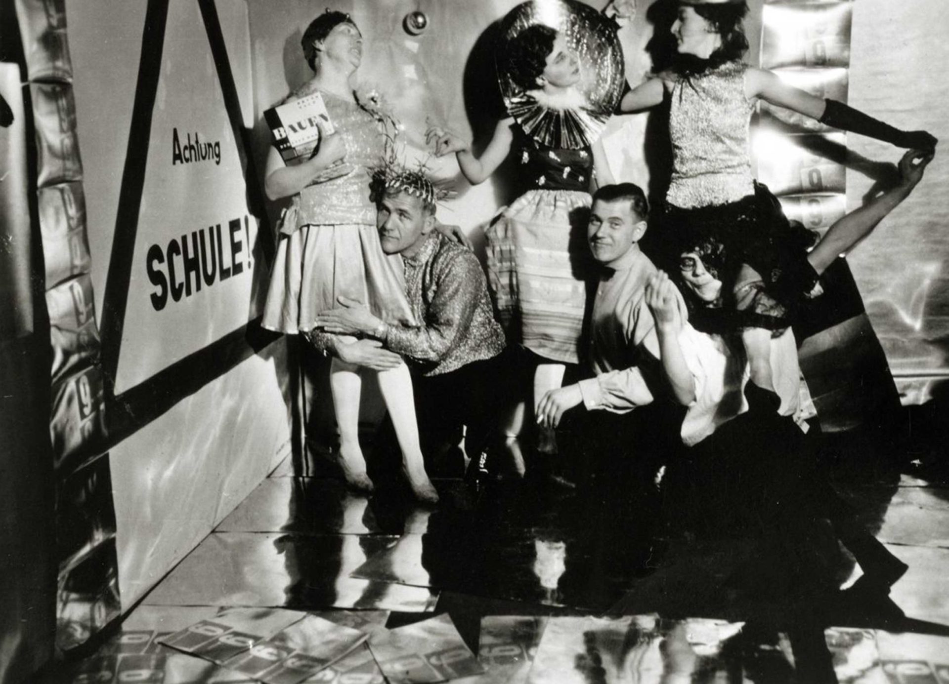 Eine Gruppe von Menschen in festlicher Kleidung posiert vor einer Wand mit der Aufschrift „Achtung Schule!“.