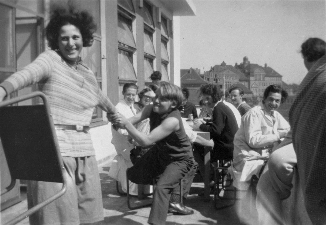 Gruppenszene mit fröhlichen Menschen beim Essen und Spielen im Freien, umgeben von Gebäuden und Fensterlicht.
