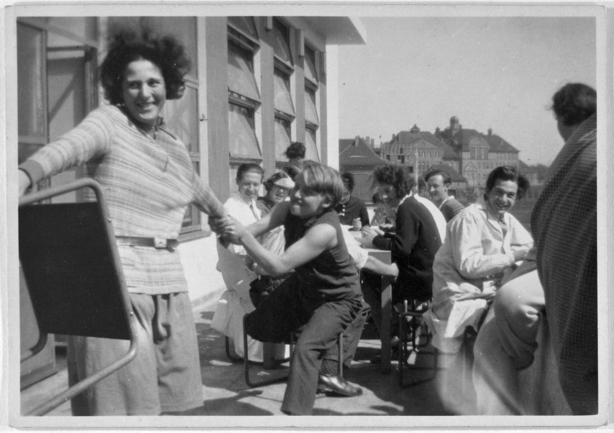 Group scene with happy people eating and playing outdoors, surrounded by buildings and sunlight.
