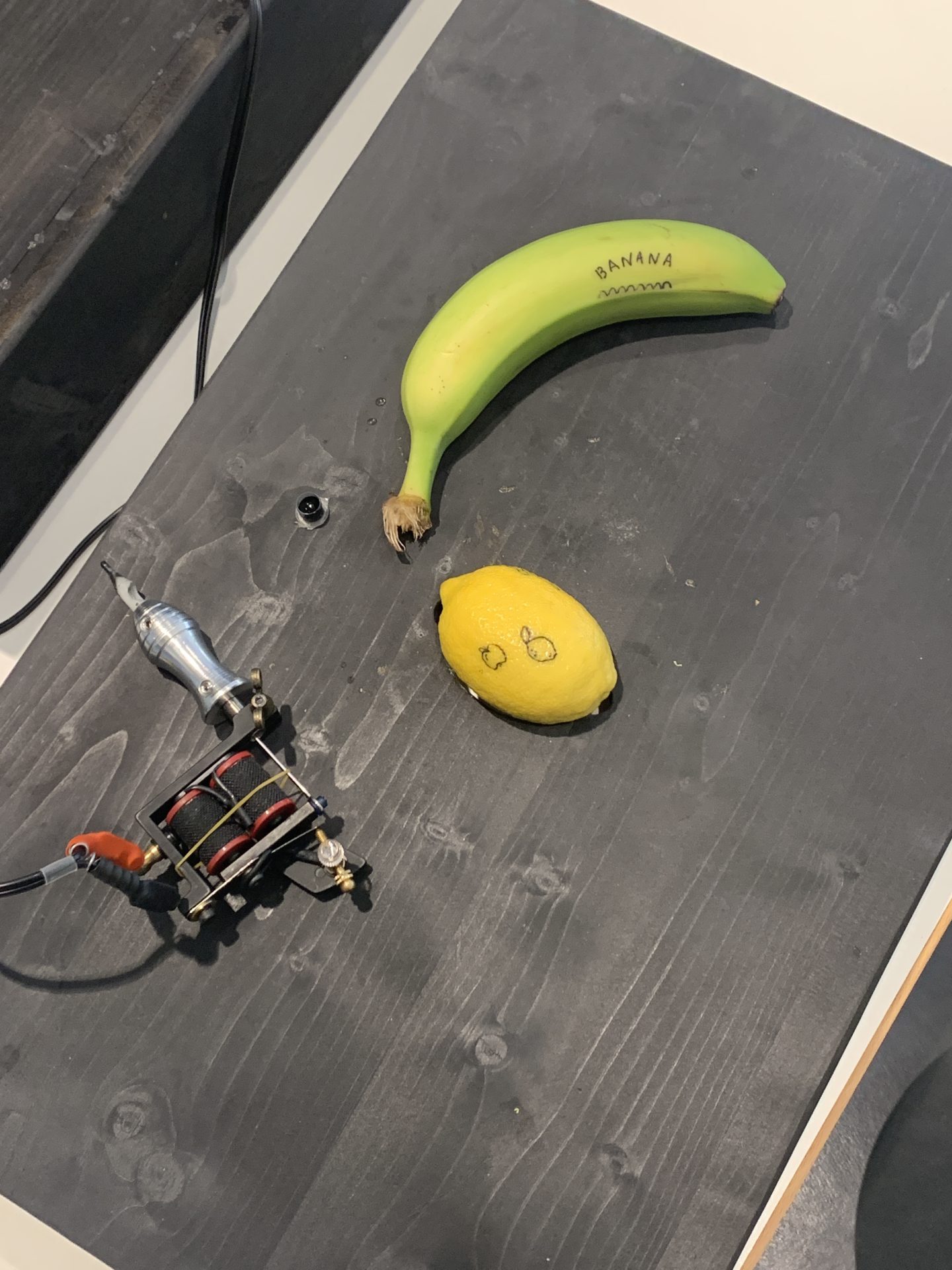 Tattoo-Maschine, Banane mit Schriftzug und gelbe Zitrone mit Gesicht auf dunklem Holztisch angeordnet.