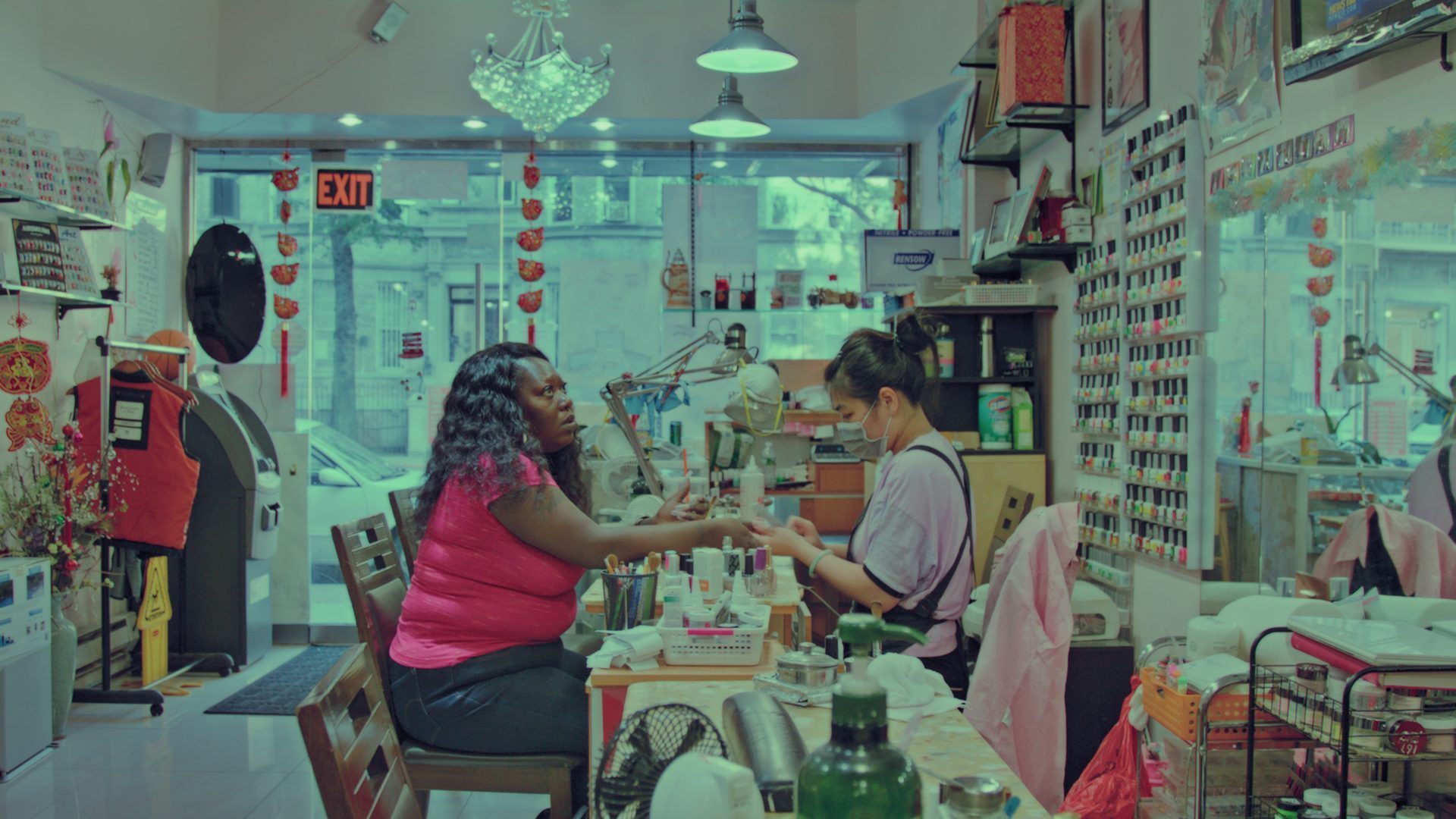 Eine Nagelpflegesitzung im Salon mit Kundin und Mitarbeiterin, umgeben von Nagellacken und Pflegeprodukten.