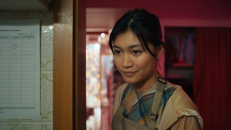 Junge Frau mit lächelndem Gesicht steht in einem warm beleuchteten Raum und schaut zur Seite.