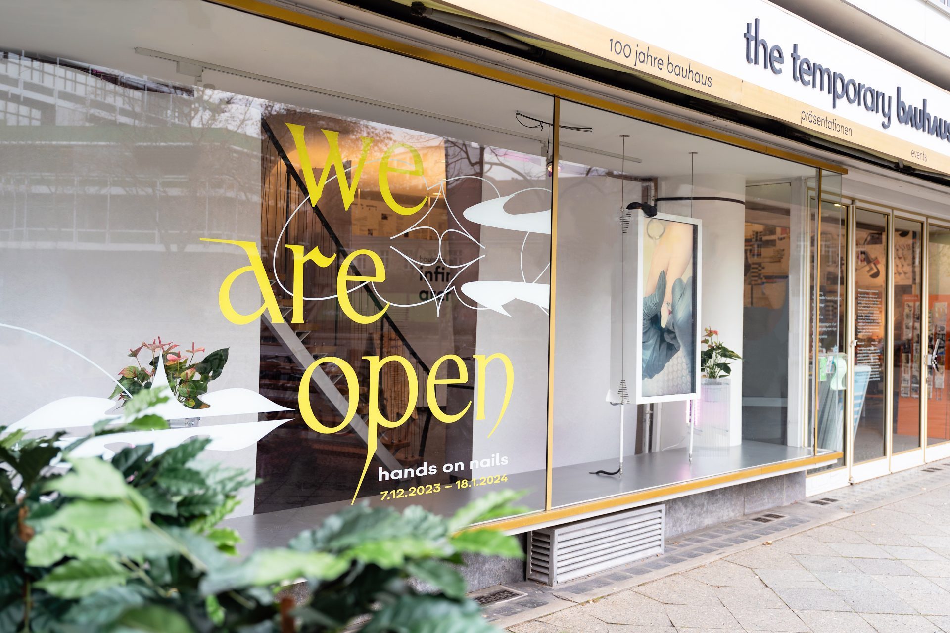 Schaufenster mit "Wir haben geöffnet"-Schild und Hinweisen zu "hands on nails". Grünes Pflanzenarrangement im Vordergrund.