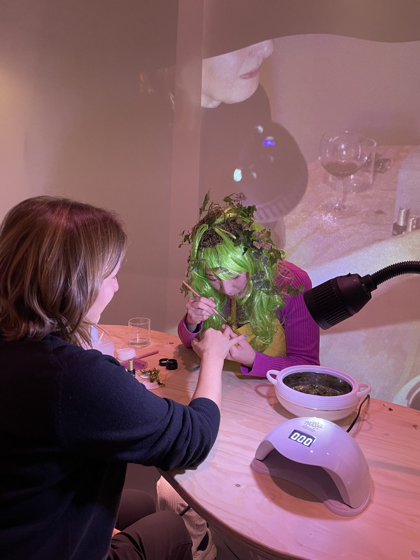 Frau mit grünen Haaren und Blattdeko bekommt eine Maniküre, während im Hintergrund eine andere Frau arbeitet.
