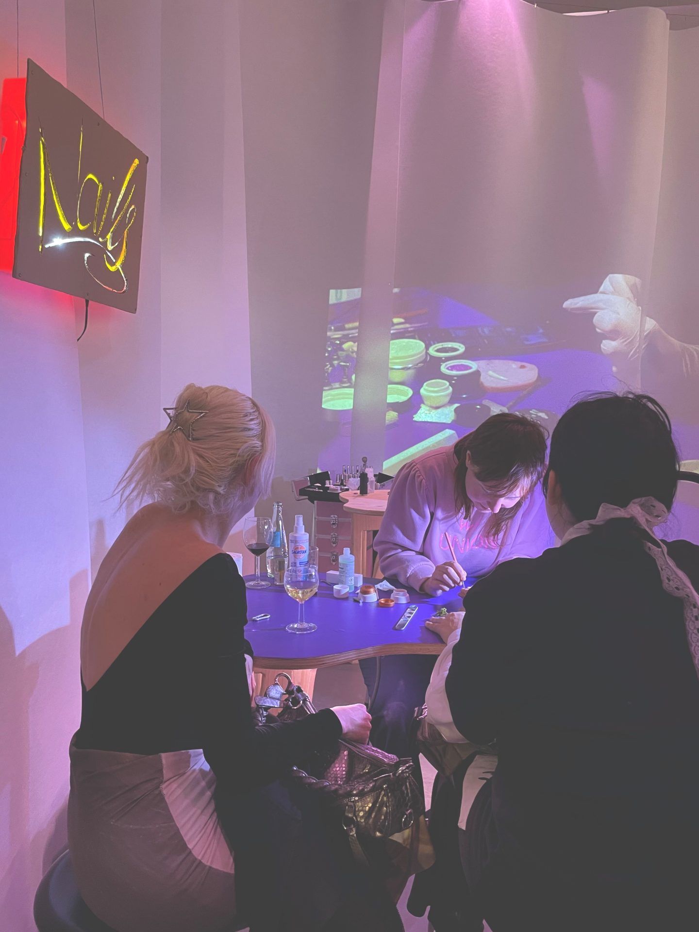 Kreatives Event mit zwei Gästen, die einem Künstler am Tisch zuschauen; im Hintergrund leuchten Projektionslichter.