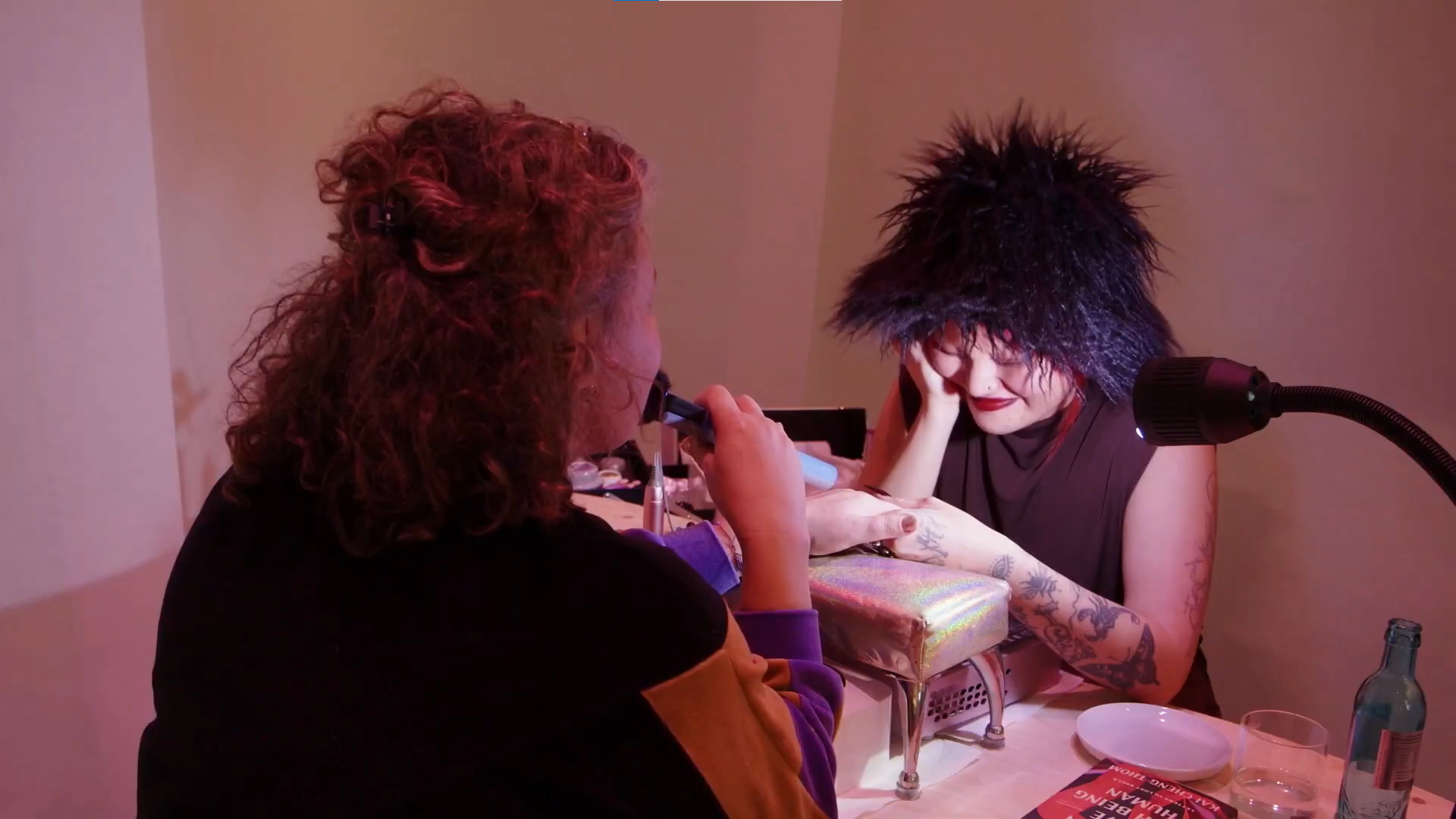Zwei Personen bei einer Maniküre, eine trägt einen auffälligen Hut und Tattoos, das Licht schafft eine besondere Atmosphäre.