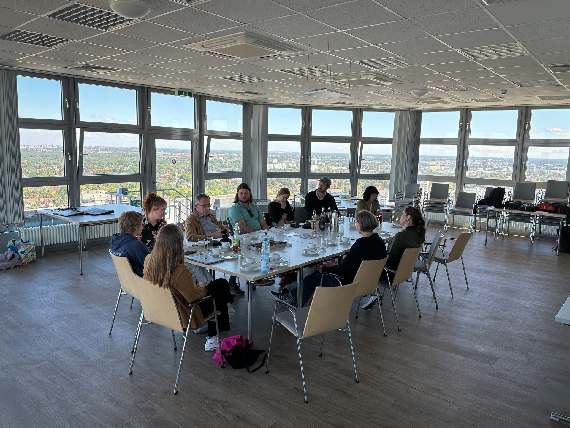 Gruppentreffen in einem modernen Raum mit großen Fenstern und Blick auf die Stadt. Teilnehmer sitzen um einen Tisch.