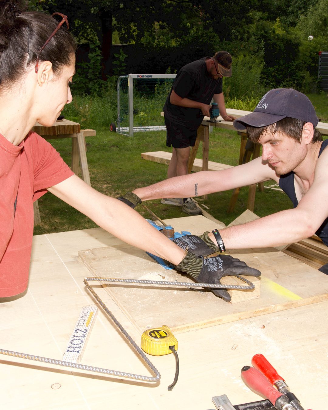 Drei Personen arbeiten im Freien an einem Holzprojekt, nutzen Werkzeuge und messen Materialien gemeinsam.