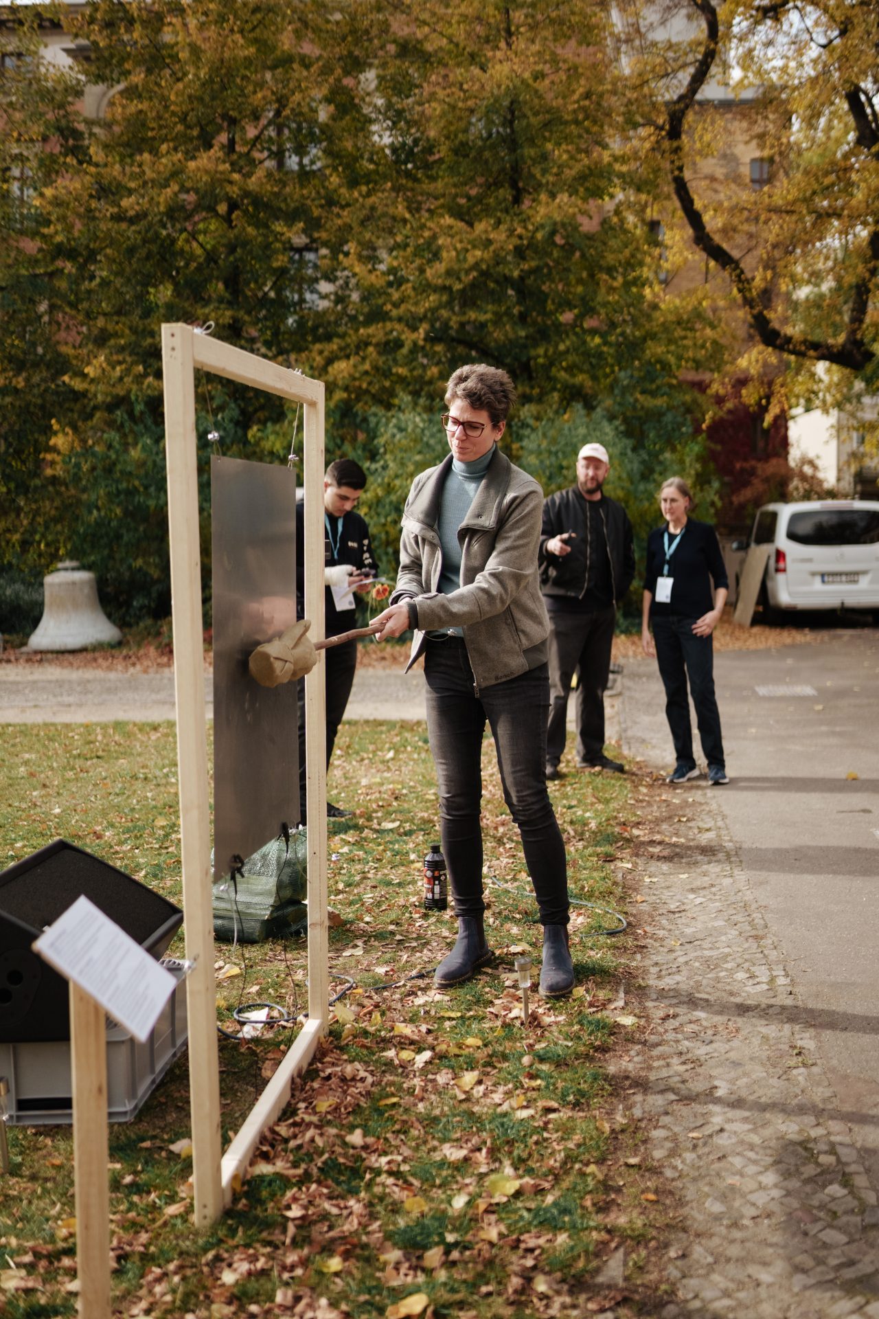 A person strikes a metal object with a tool while spectators stand in the background. Autumn setting.