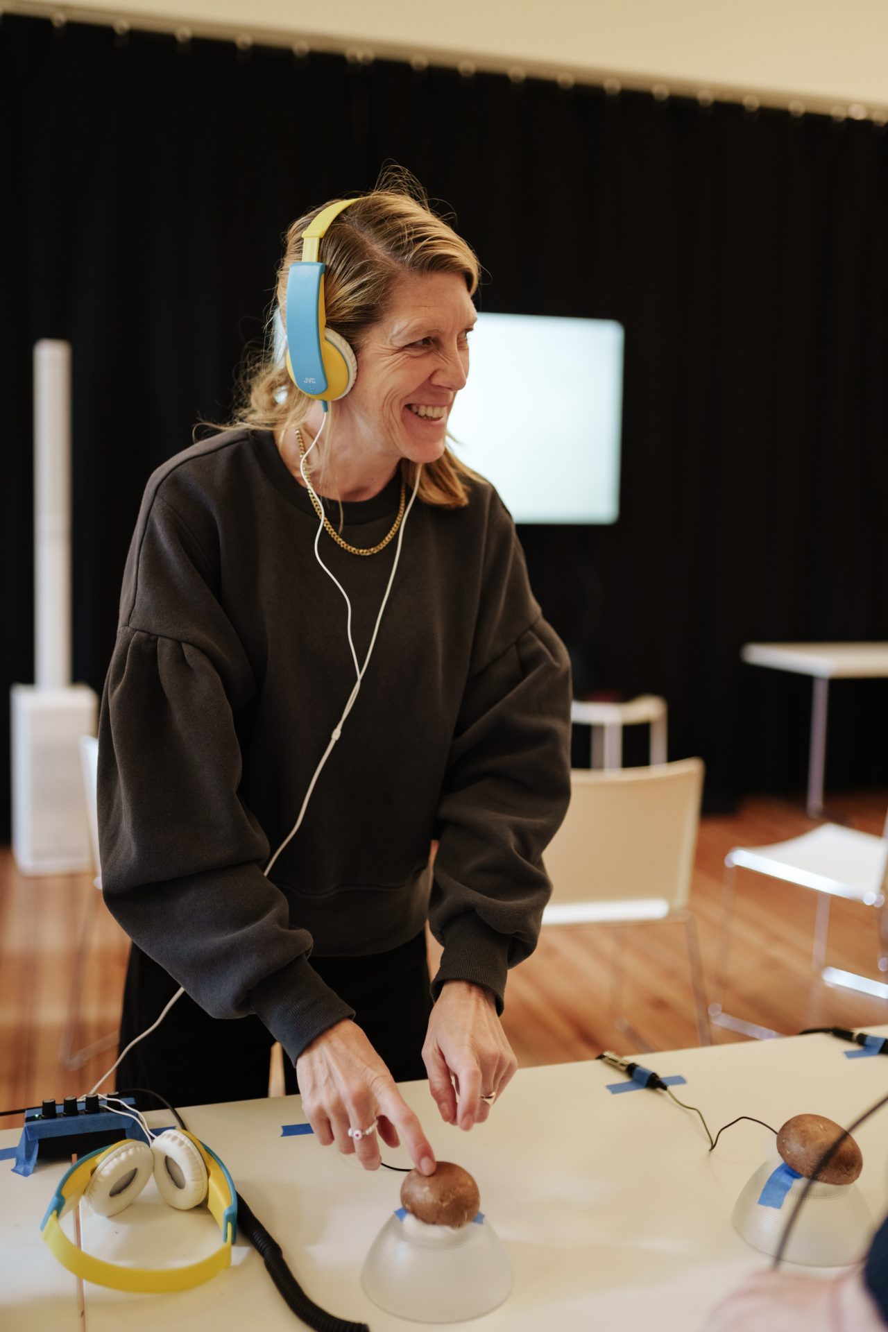 Frau mit Kopfhörern, die an einem Tisch mit Geräten experimentiert und dabei lächelt.